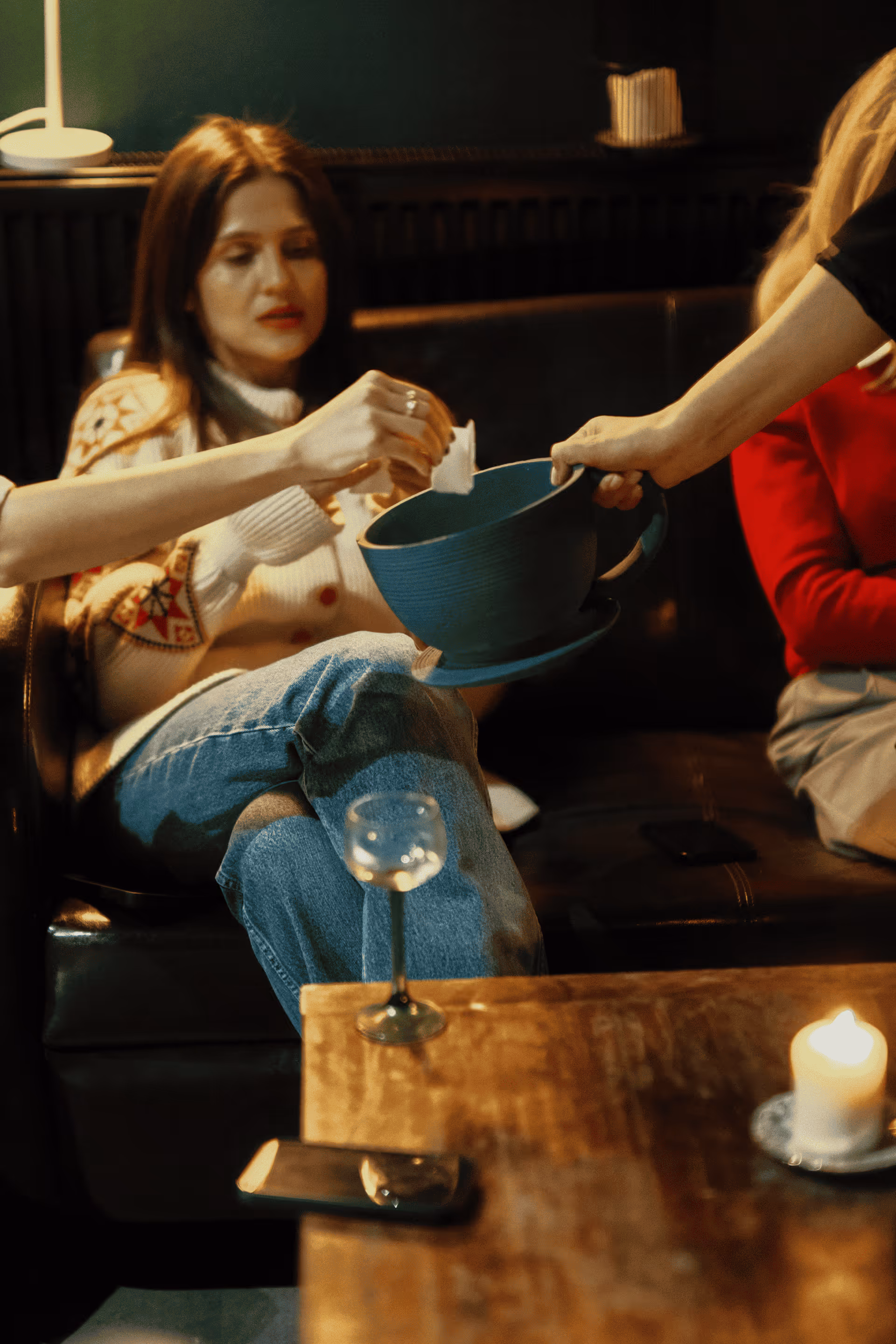 People sitting on a sofa passing a large blue cup while a glass of water and a lit candle are on a wooden table in the foreground.