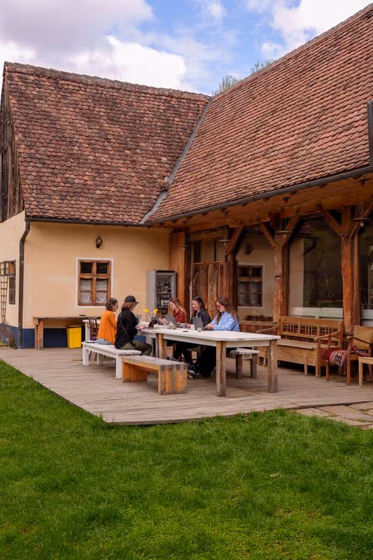 Five people sitting around a wooden outdoor table on a patio adjacent to a rustic house with a tiled roof and wooden beams.