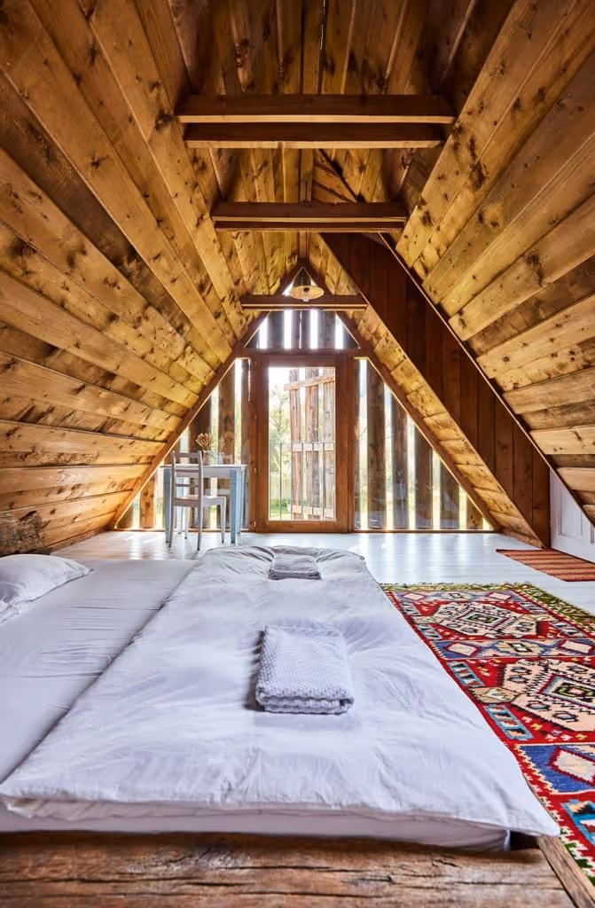 Cozy A-frame cabin bedroom with low bed, white bedding, colorful rug, wooden walls, and small table by a glass door.