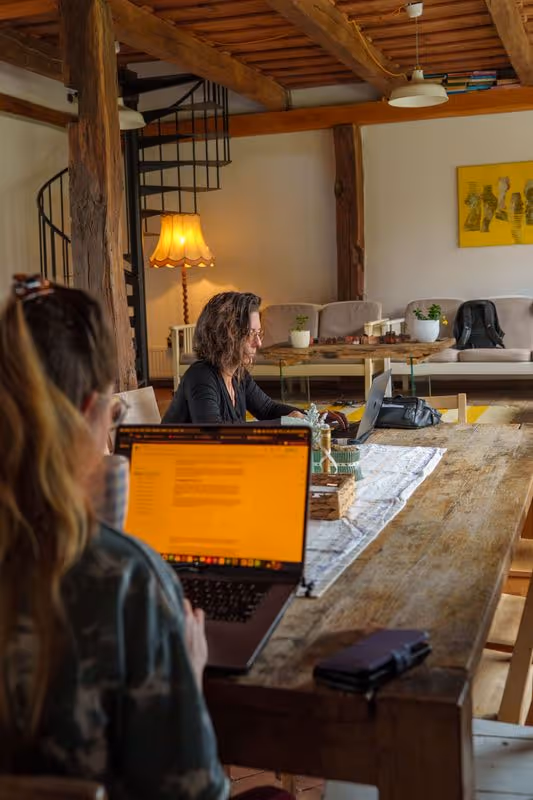 Two women working on laptops in a cozy room with wooden beams, a spiral staircase, and a couch with plants and a yellow painting on the wall.
