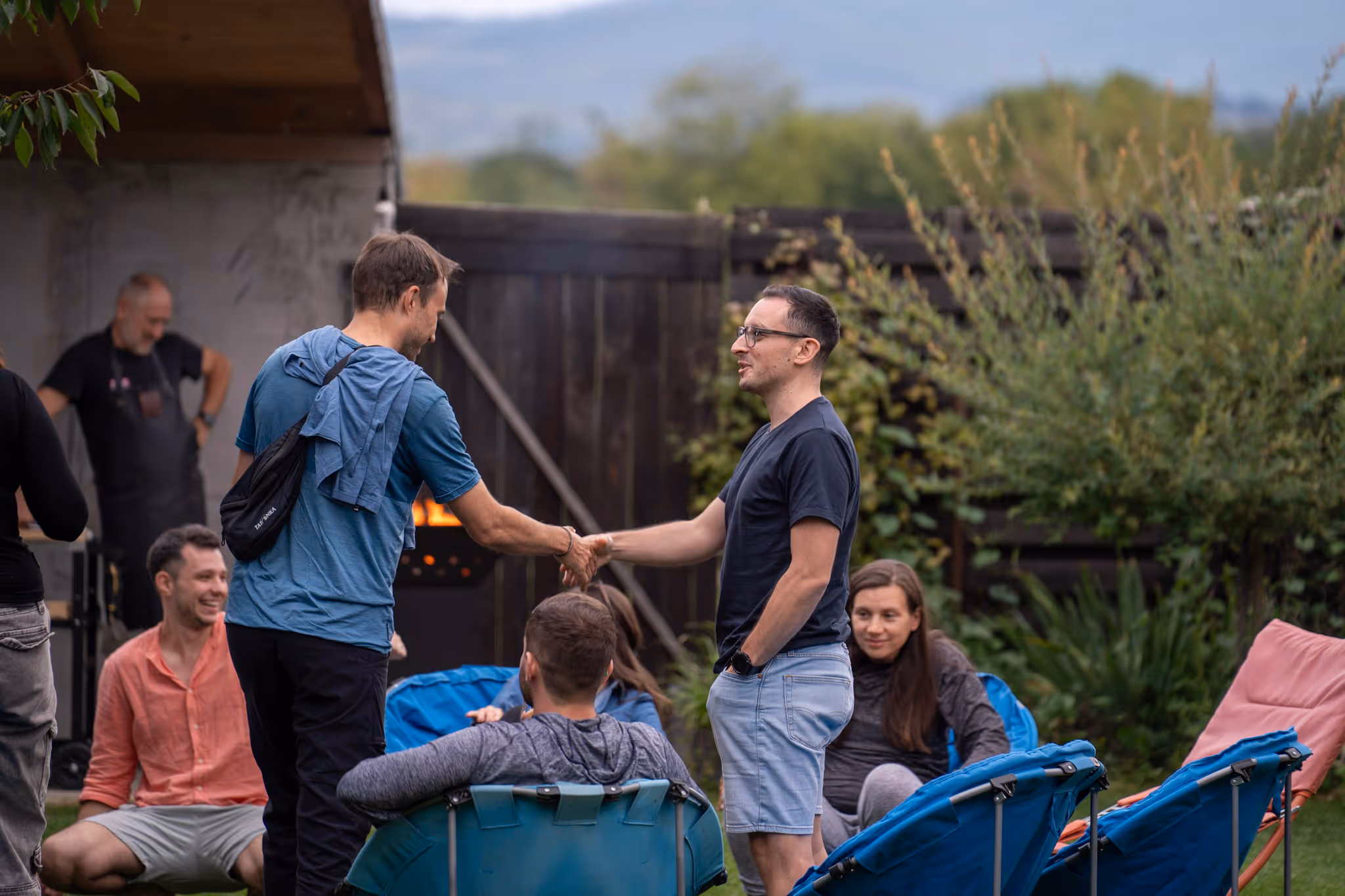 Two men shaking hands at an outdoor gathering with others sitting on chairs and a grilling area in the background.