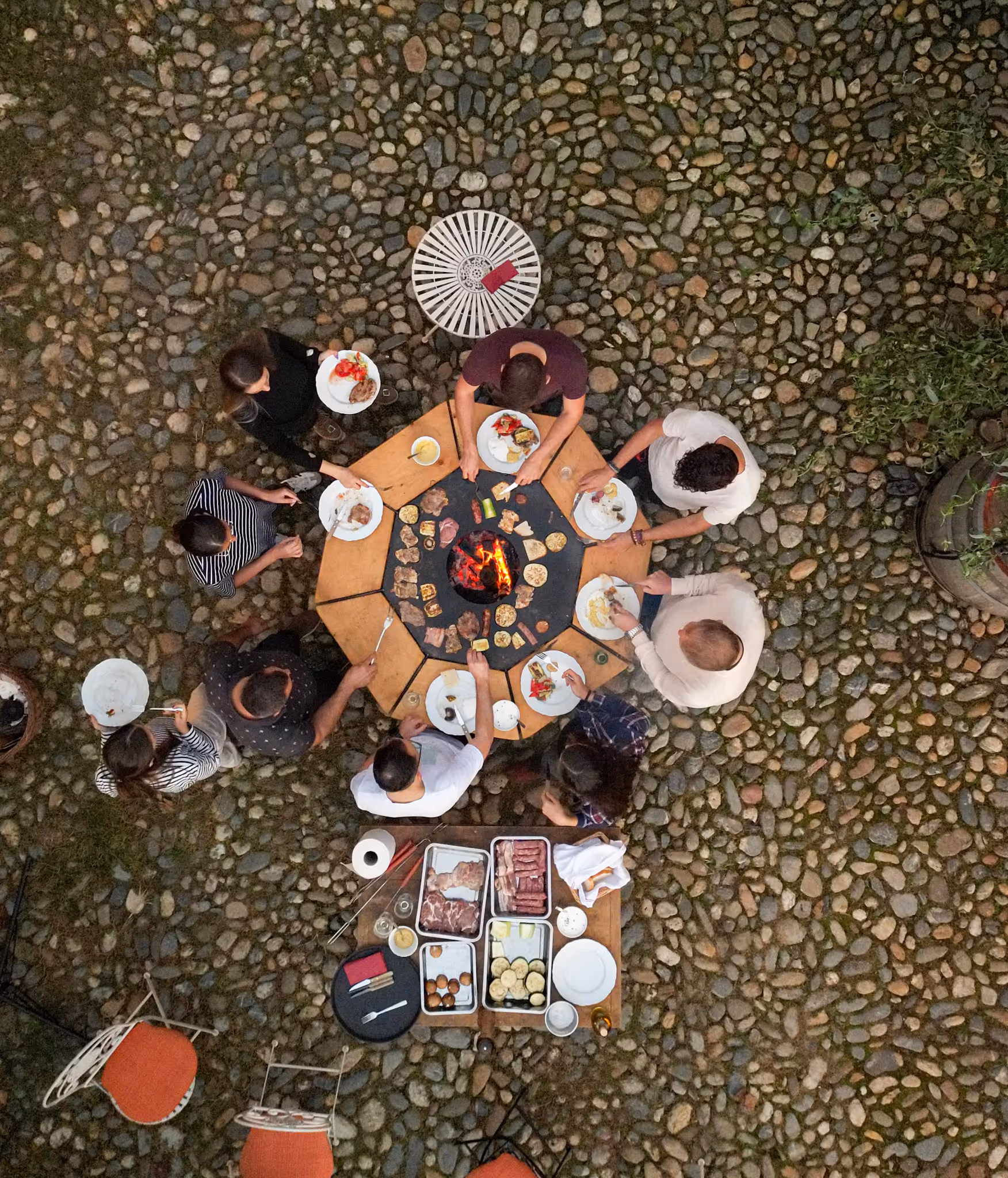 Group of people gathered around an outdoor round grill table cooking meat and vegetables on a cobblestone patio.