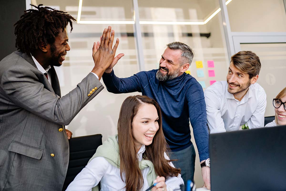 A group of people smiling around a desk while two men give each other a high five.