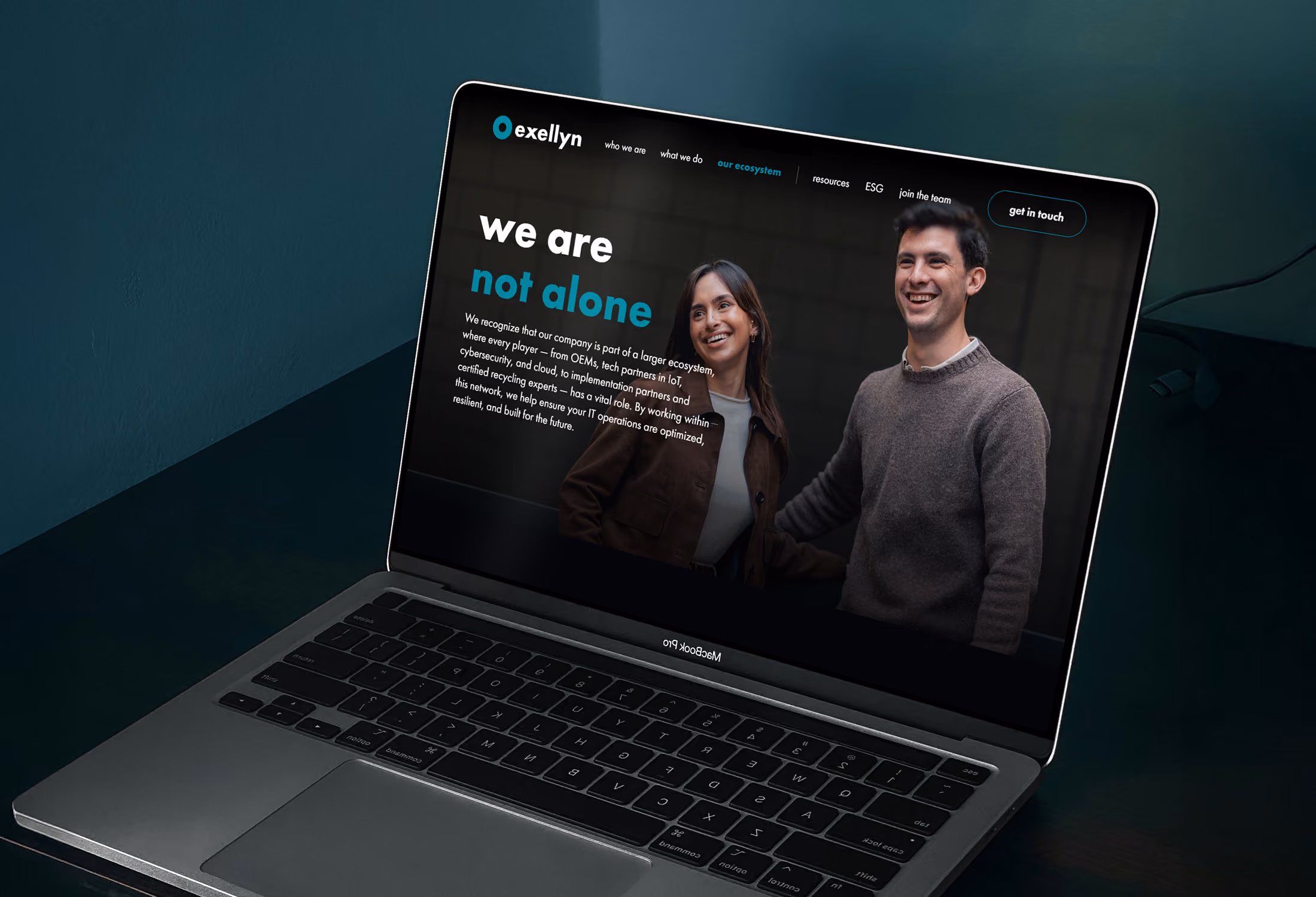 Open MacBook Pro laptop on a dark surface displays a website with the header 'we are not alone' featuring a smiling woman and man standing side by side, with text describing company collaboration and ecosystem partnerships.