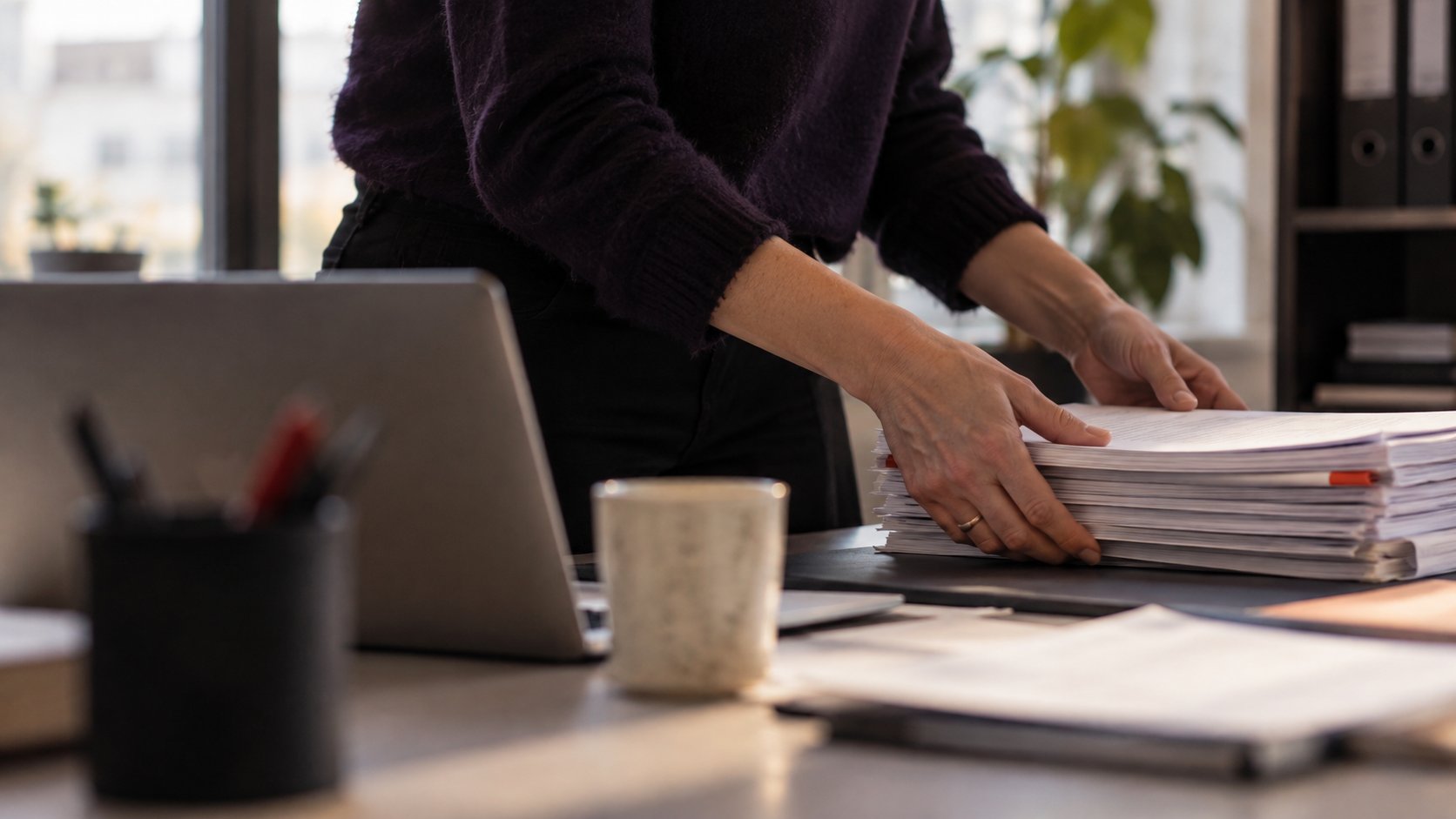 Handen leggen een stapel papieren documenten naast een laptop