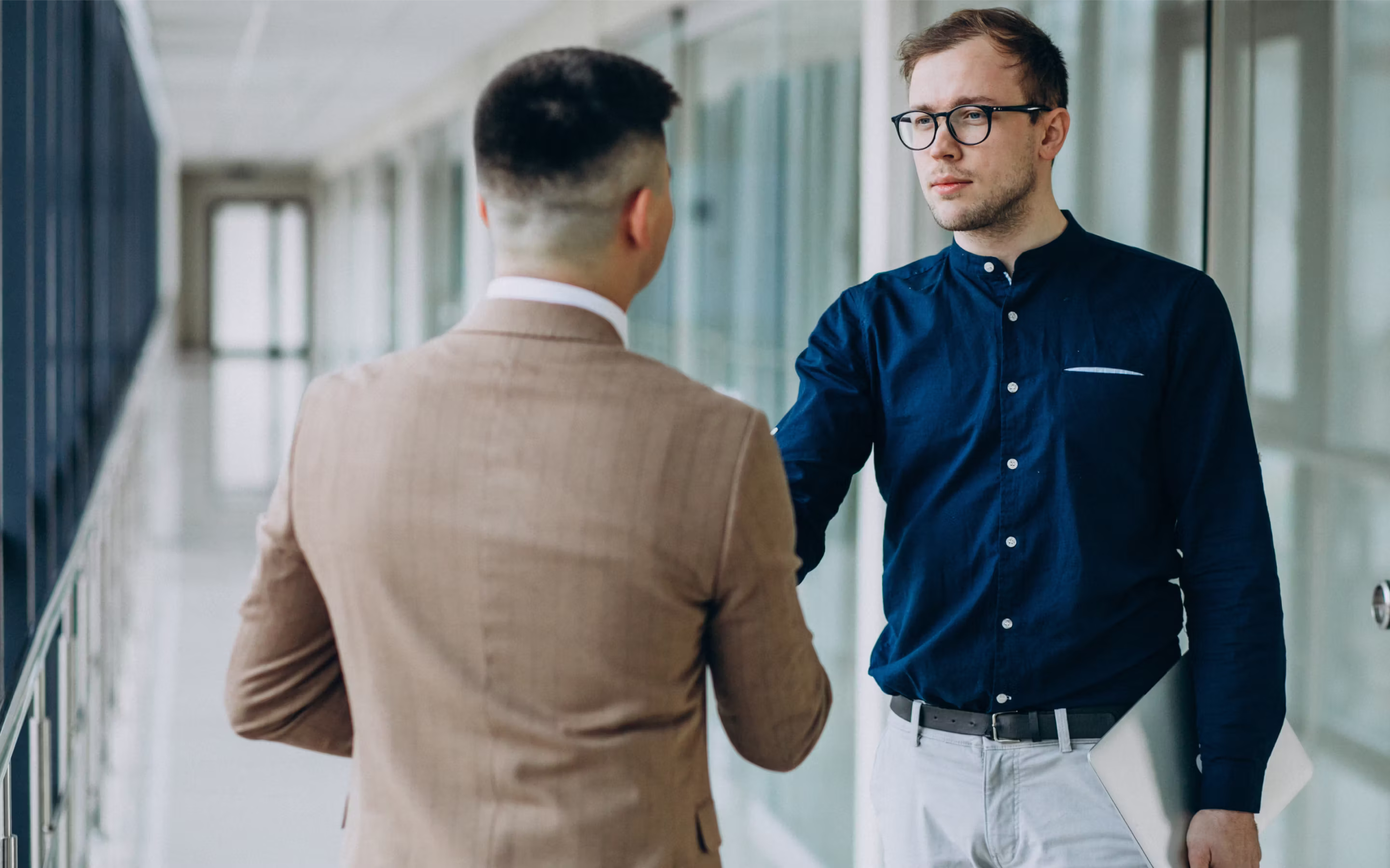 Two men having a professional conversation in a modern office hallway, one holding a laptop.