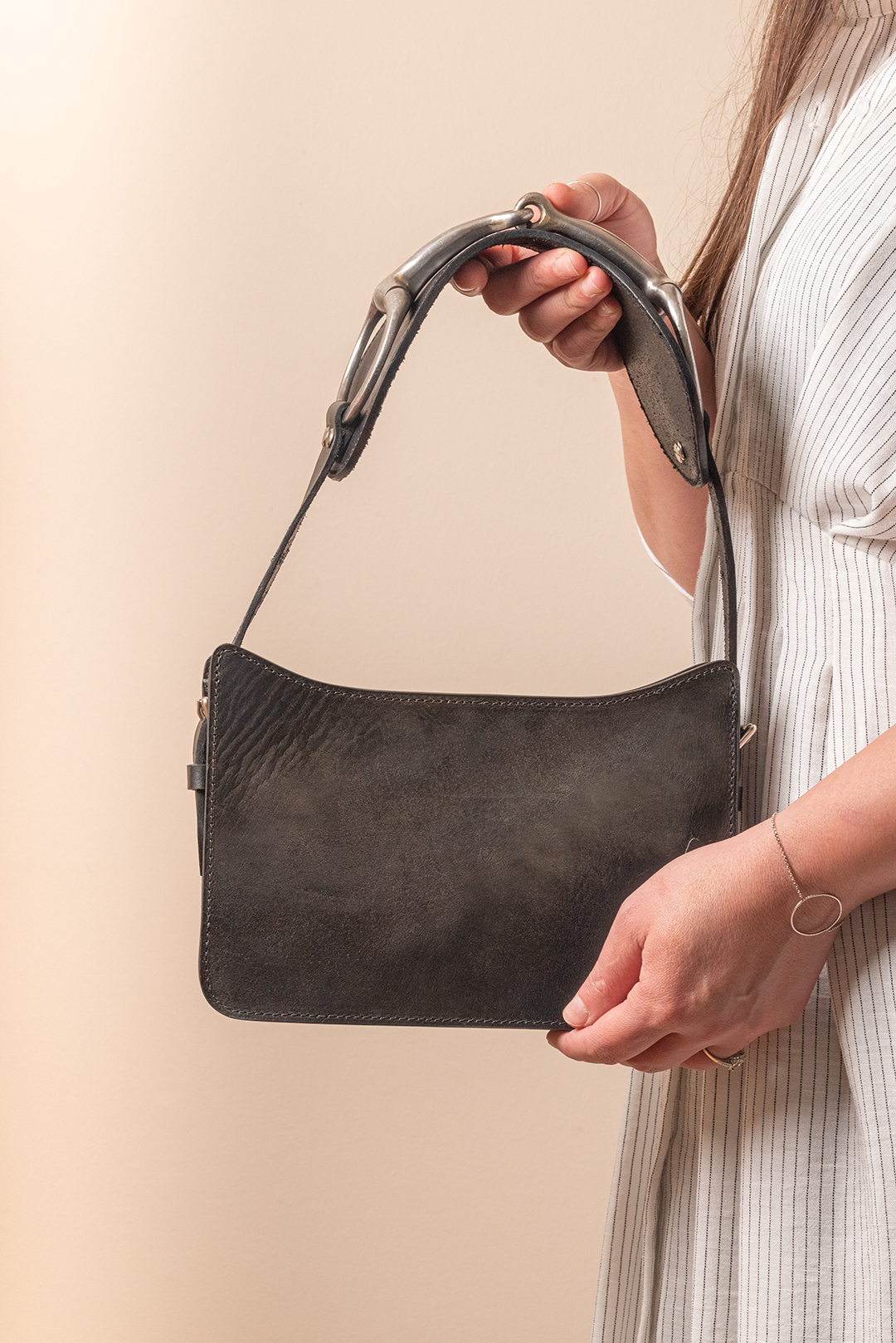 Person holding a black leather handbag with a short strap and metal hardware against a beige background.