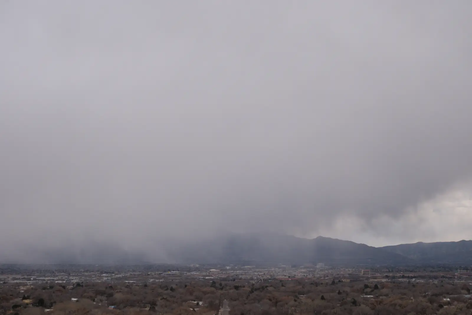 Dark winter storm clouds and falling snow over Albuquerque, New Mexico, with the Sandia Mountains partially obscured in the distance.