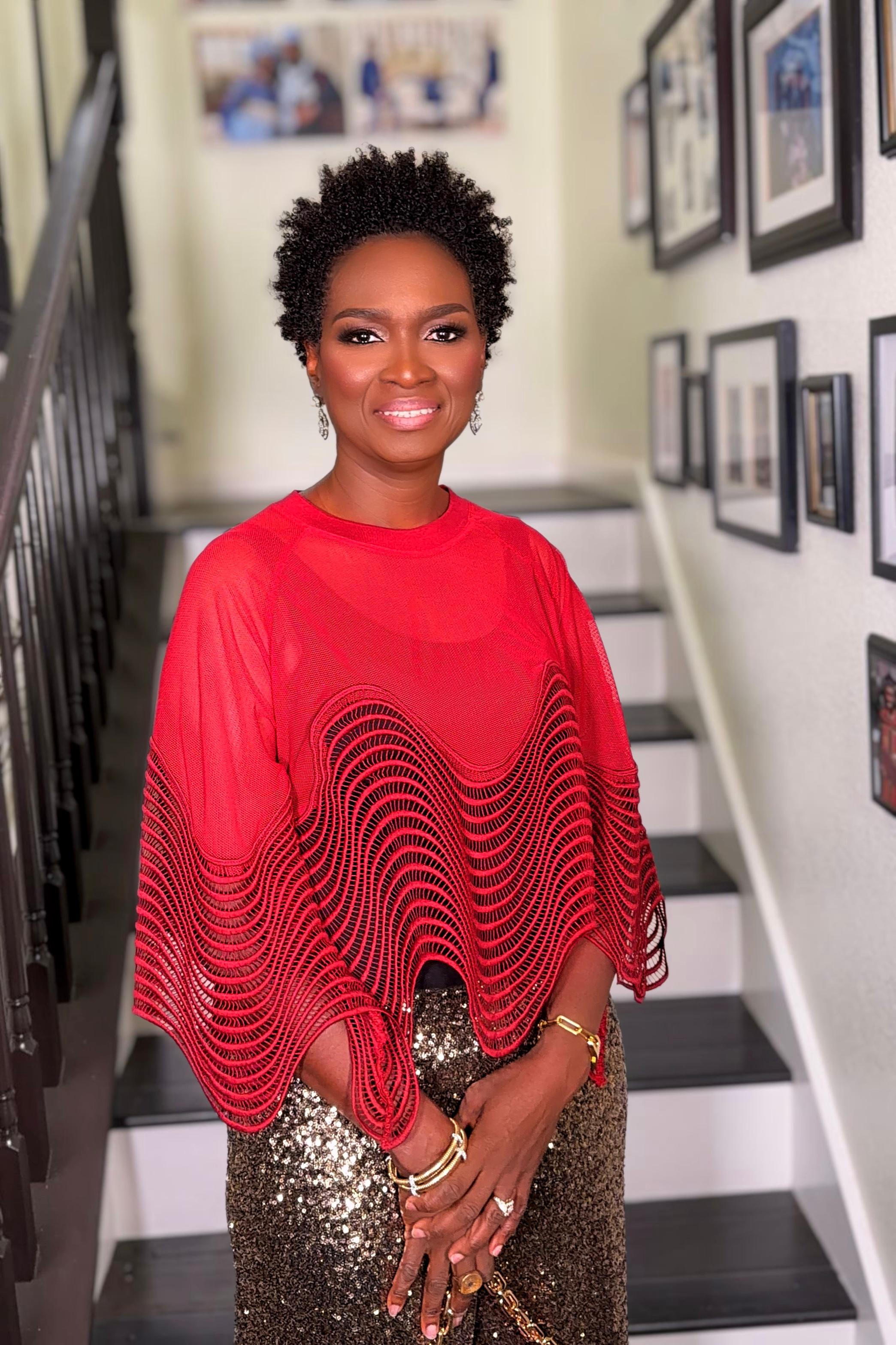 Woman with short curly hair wearing a red patterned top and sequined skirt standing on stairs with framed pictures on the wall.