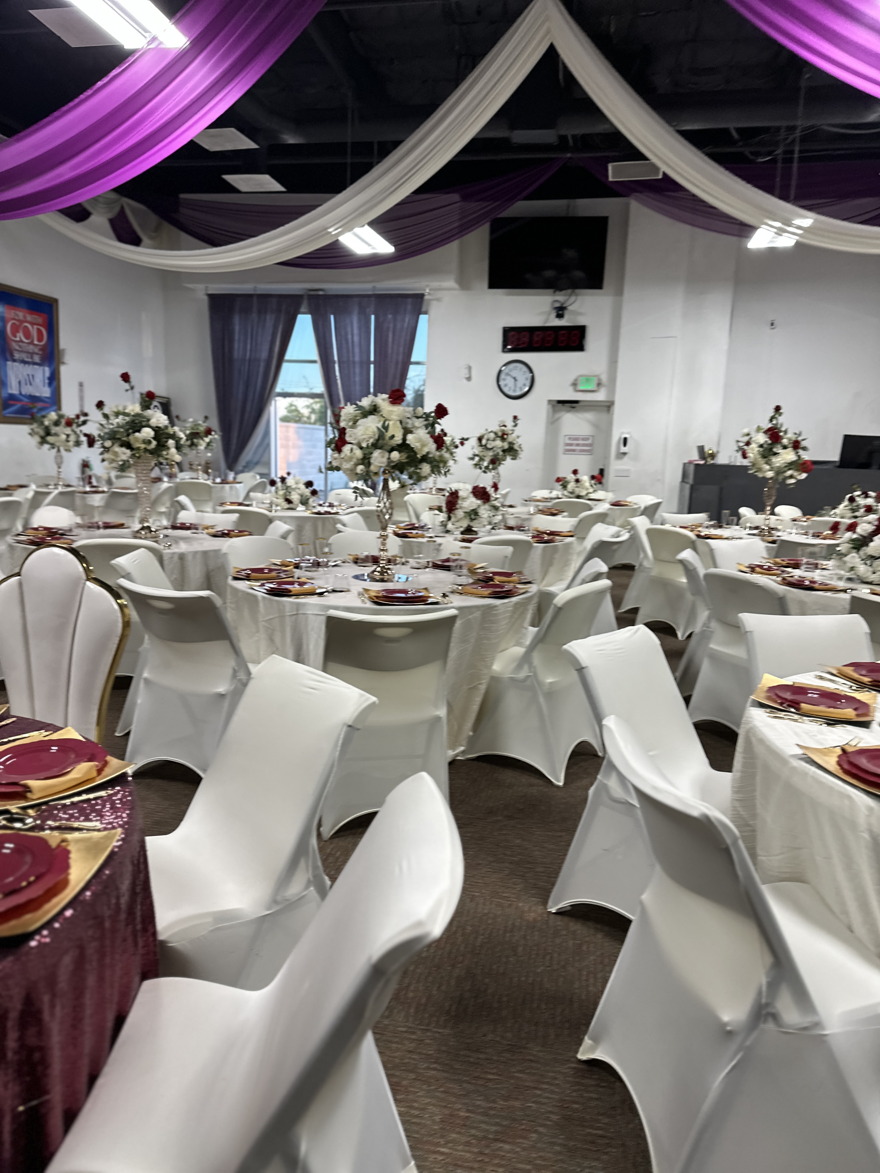 Event hall with round tables covered in white cloths, white chairs with covers, floral centerpieces, and purple and white draped ceiling decorations.