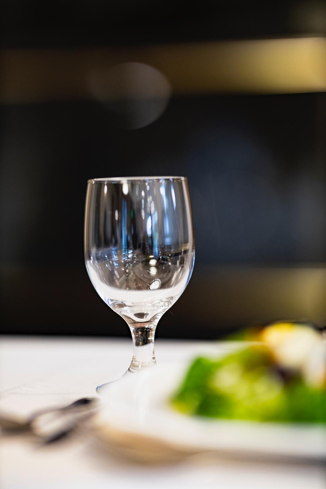 Empty clear wine glass on a white tablecloth with a blurred plate of salad and utensils in the foreground.