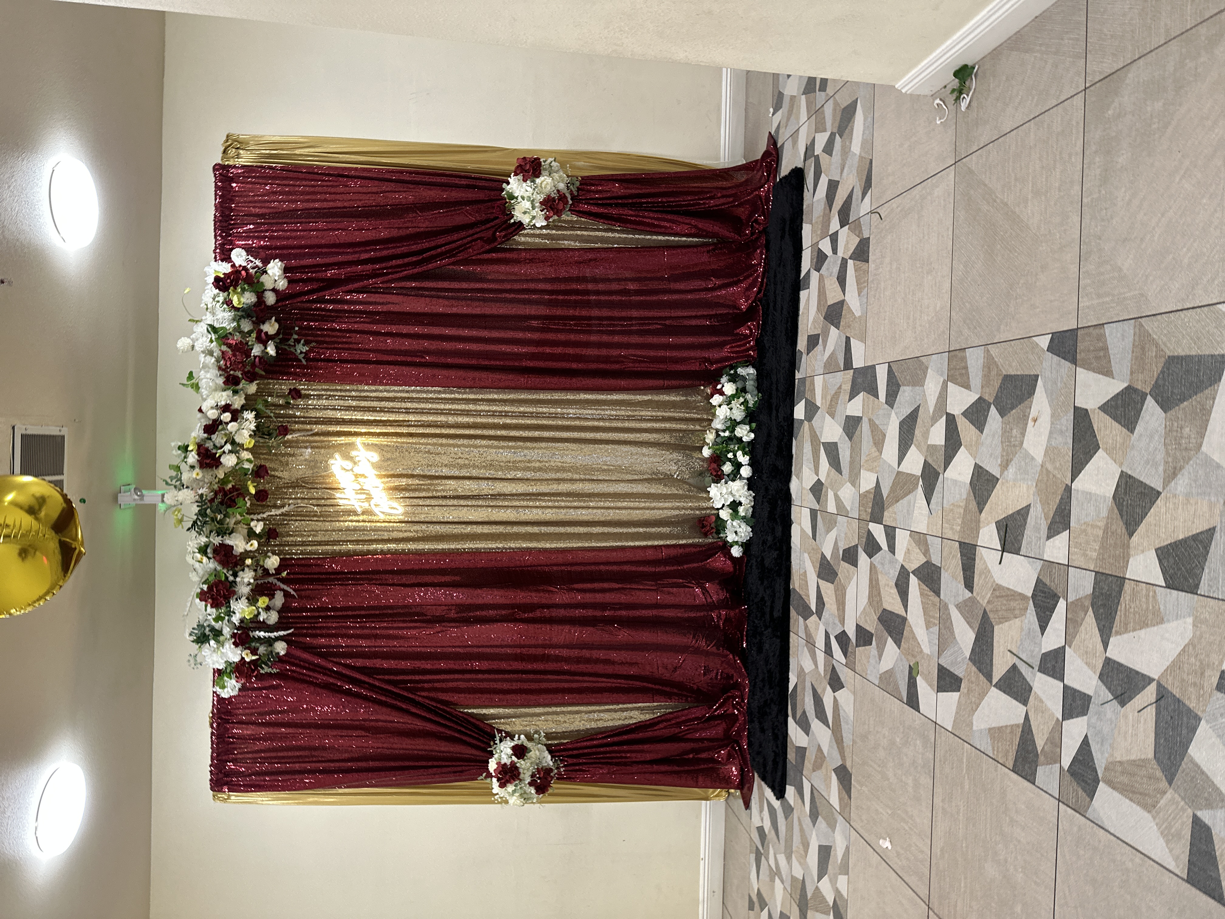 Elegant birthday backdrop with gold and burgundy shimmering curtains, floral arrangements, and a glowing Happy Birthday sign.
