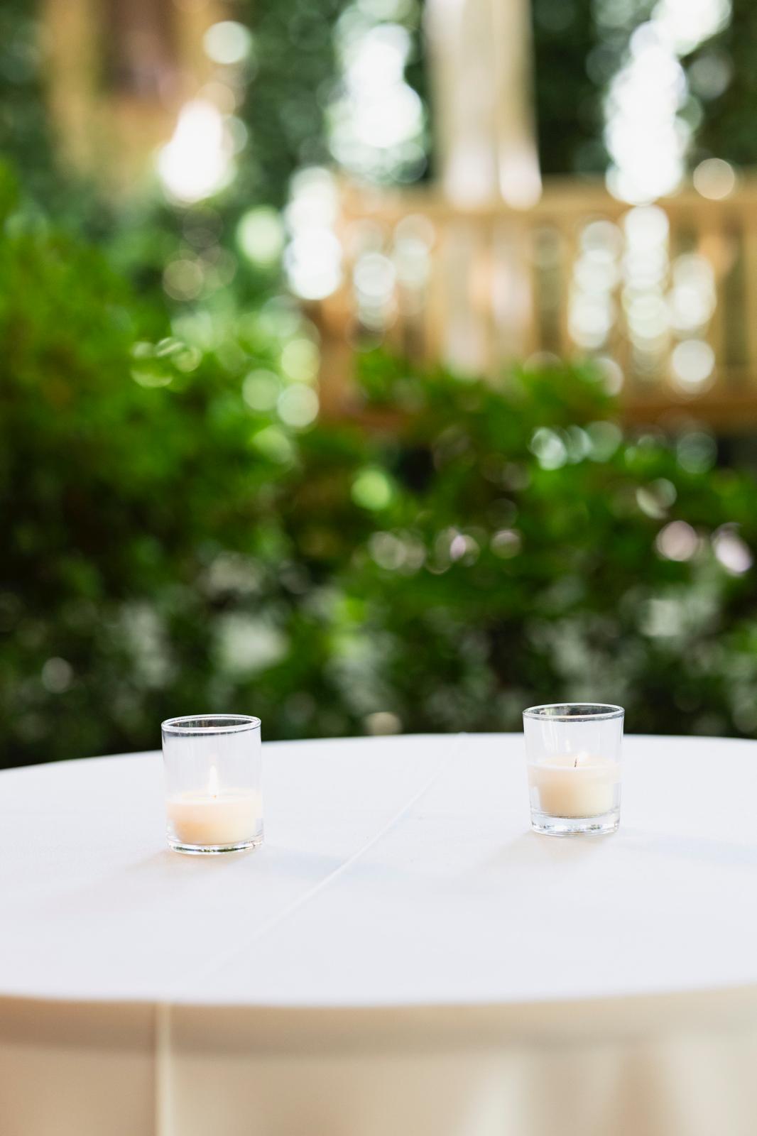 Two small glass votive candles lit on a round white tablecloth-covered table with blurred green foliage background.