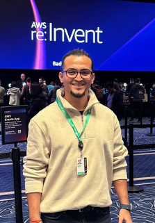 A man with glasses and a light-colored sweater smiling in front of an AWS re:Invent event screen.