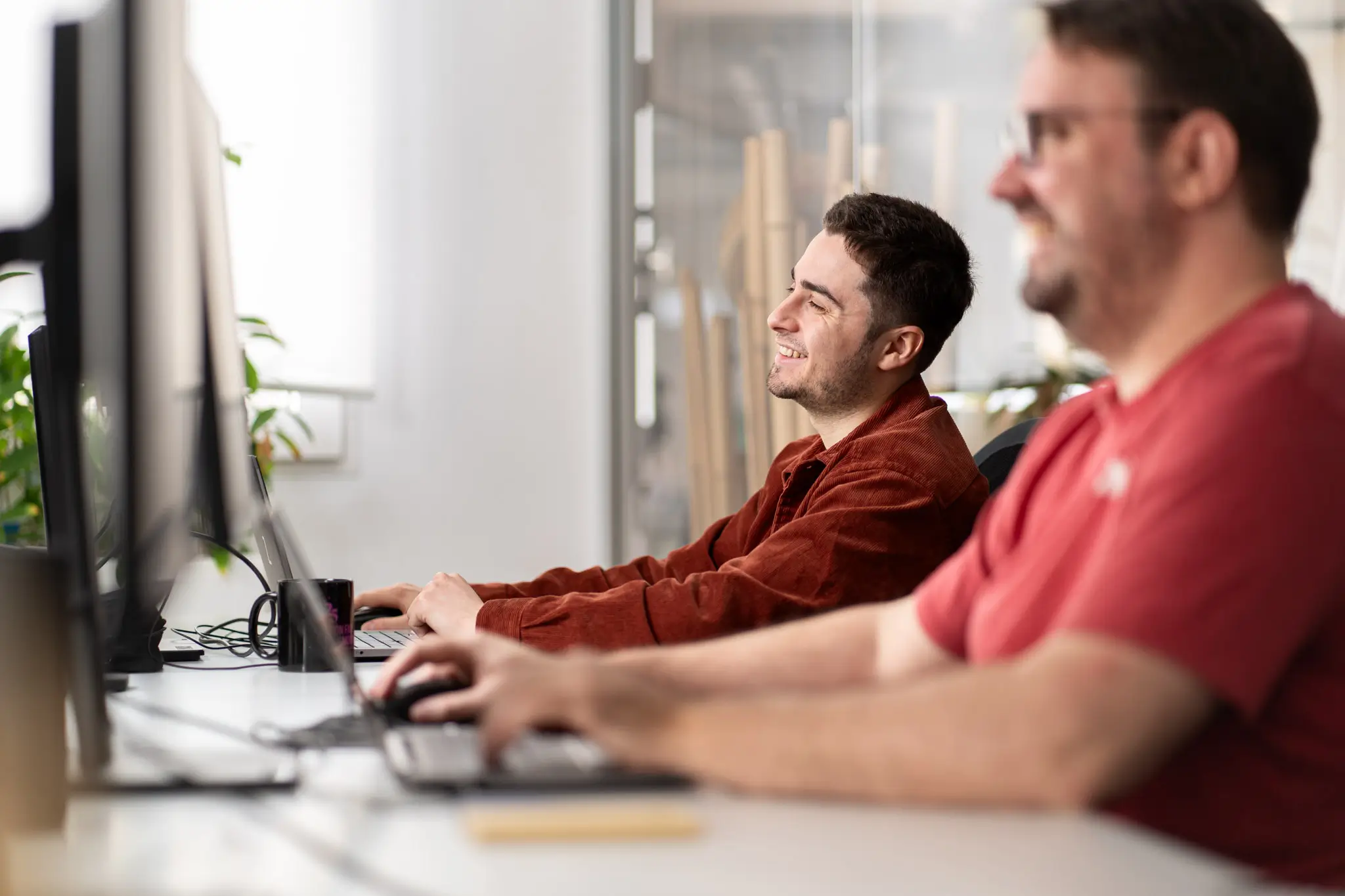 Deux hommes assis côte à côte sourient en travaillant sur des ordinateurs dans un bureau lumineux.