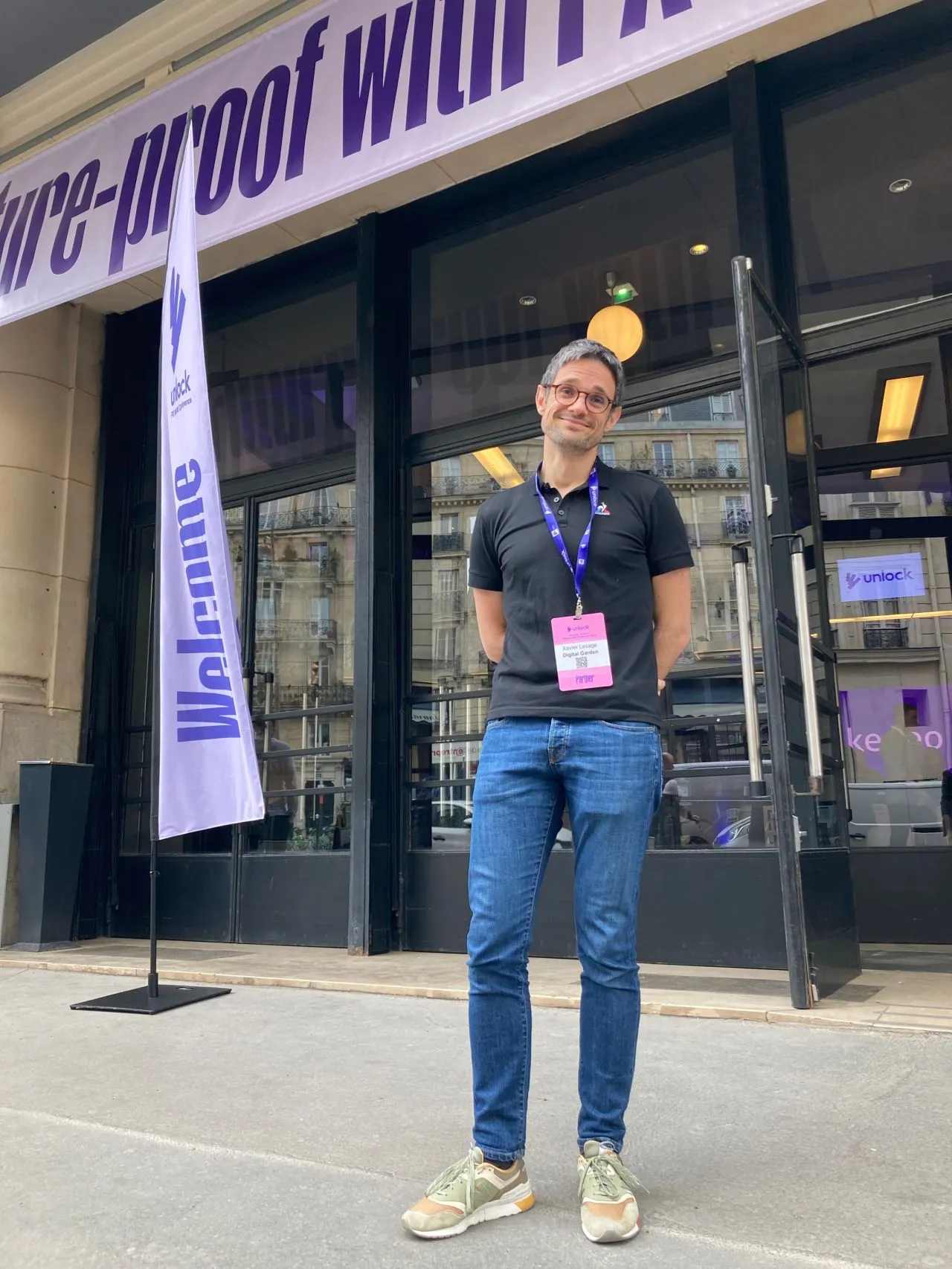 Un homme avec un badge debout devant une entrée vitrée avec une bannière Welcome à côté.