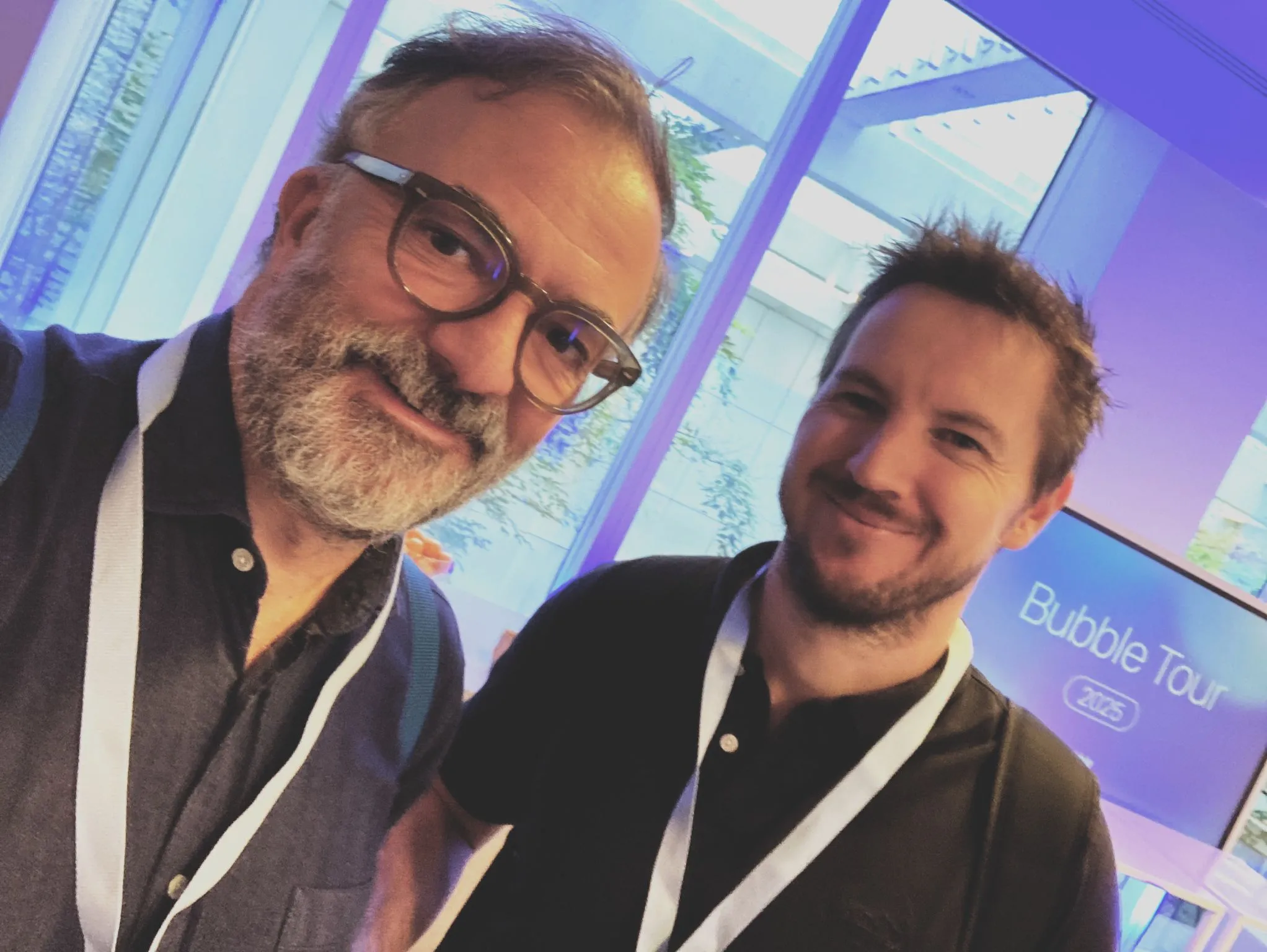 Deux hommes souriants portant des badges blancs dans un cadre intérieur moderne.
