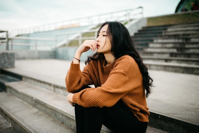 A woman in an orange sweater sitting thoughtfully on outdoor steps.