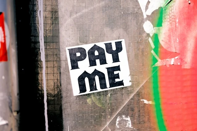 A sticker reading Pay Me on a weathered surface.