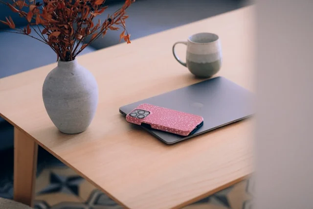 A phone, laptop, mug and vase left on a table.