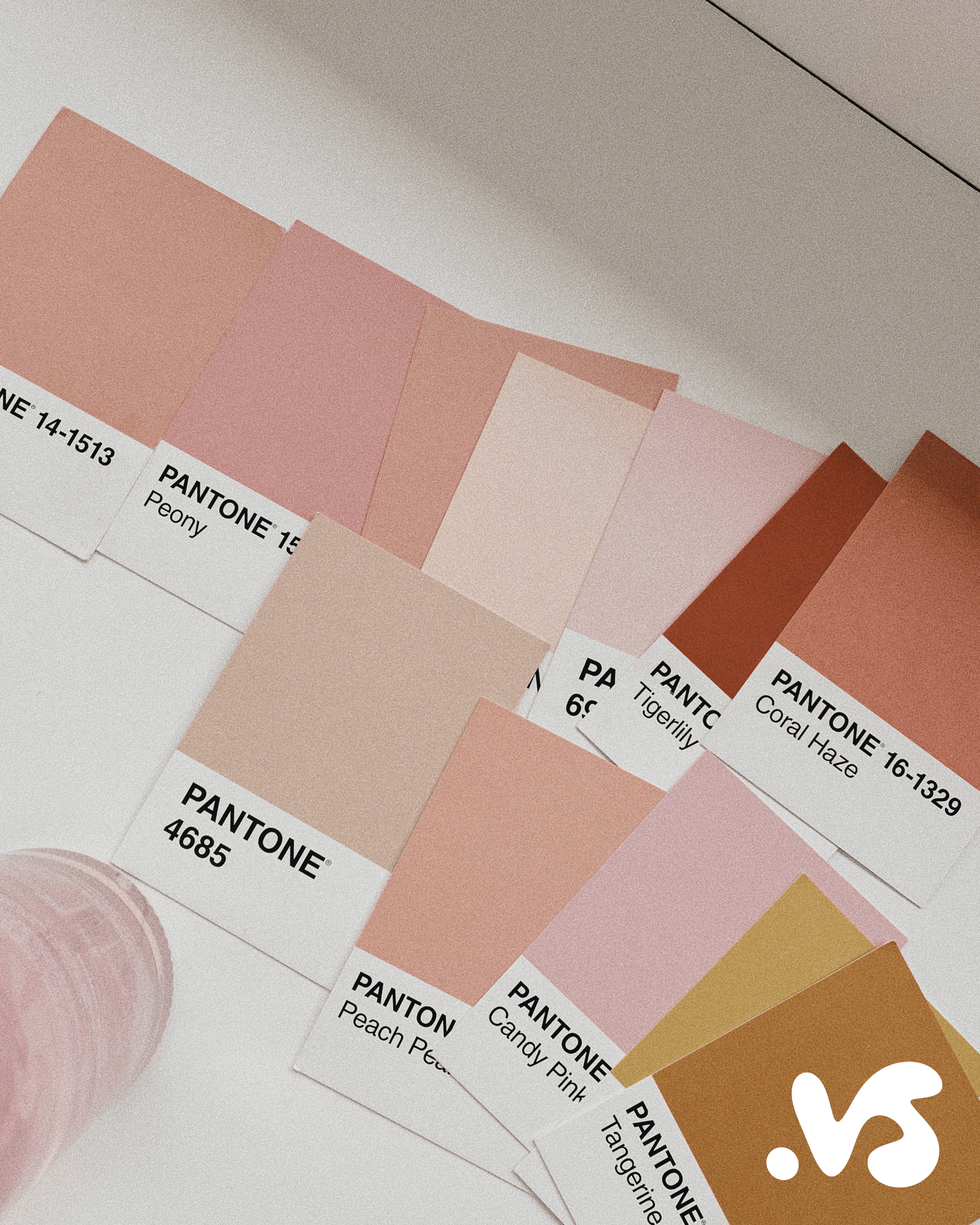 Flere Pantone-fargeprøver i nyanser av rosa, fersken og korall ligger spredt ut på en hvit overflate. Fargeprøvene er merket med fargenavn og koder. Et rosa, gjennomskinnelig objekt er delvis synlig i hjørnet.