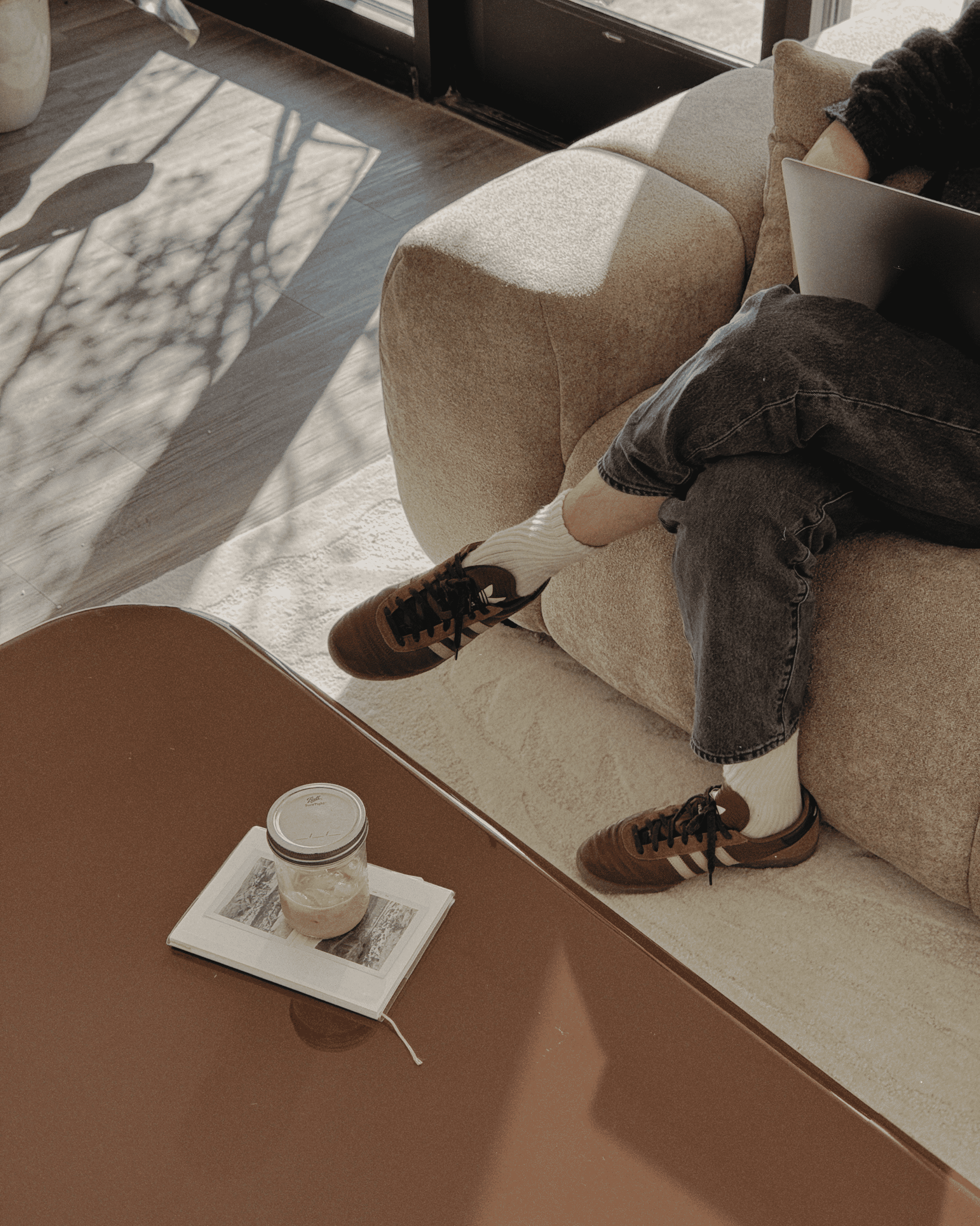 En person sitter på en beige sofa med en bærbar PC, med beina i kryss. Like ved står et brunt bord med en notatbok, en penn og en krukke med iskaffe. Sollyset kaster skygger over scenen.
