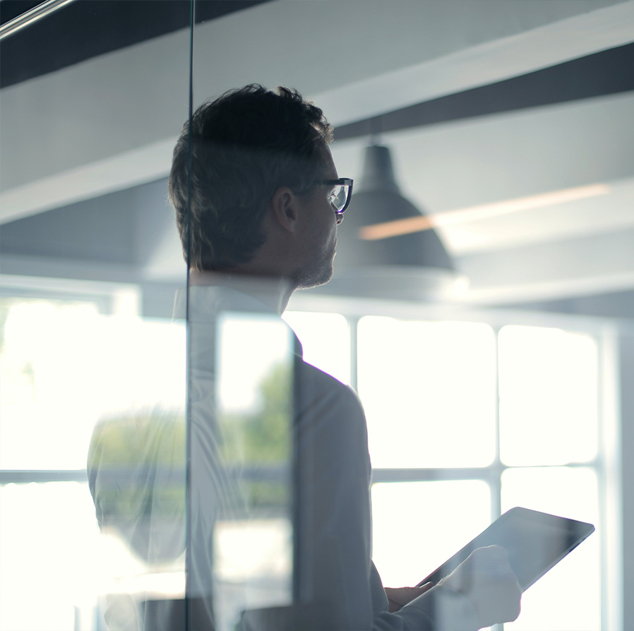 Side profile of a man wearing glasses holding a tablet, seen through a glass window in a bright modern office.