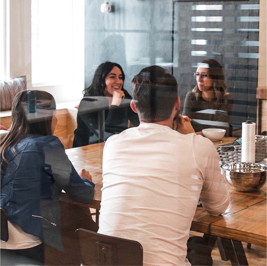 Four people sitting around a wooden table in an office, engaged in a conversation with two women smiling and talking, seen through a glass partition.