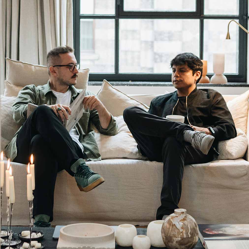 Two men sitting on a beige sofa having a conversation; one holds a newspaper and the other a cup.