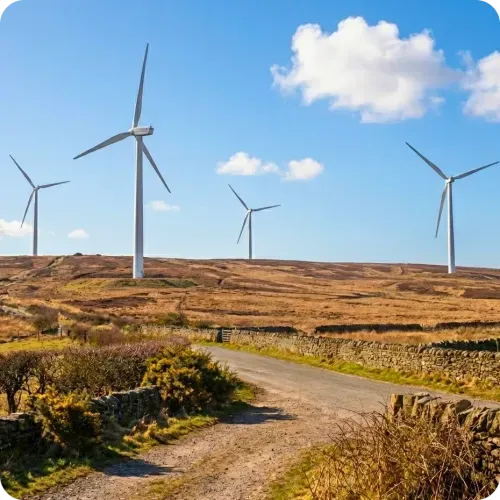 Rural landscape with a dirt road and stone fences leading to a hill dotted with four large wind turbines under a blue sky with scattered clouds.