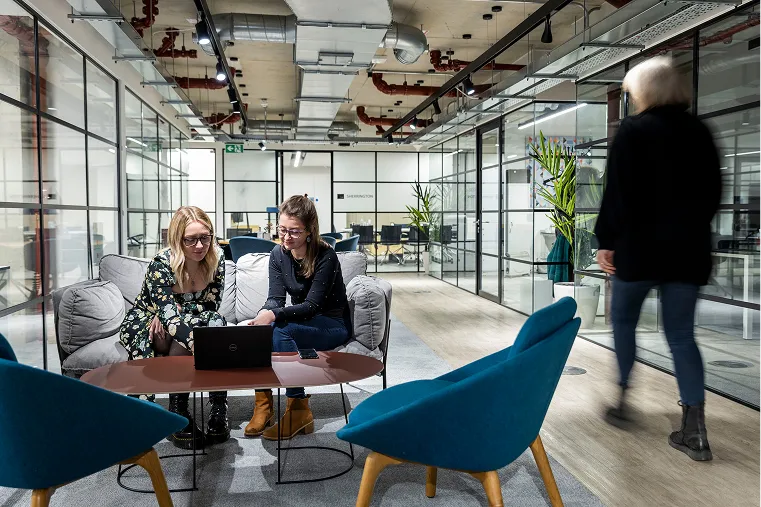 Two women working on a laptop at a low table in a modern open-plan innovation centre while a person walks past