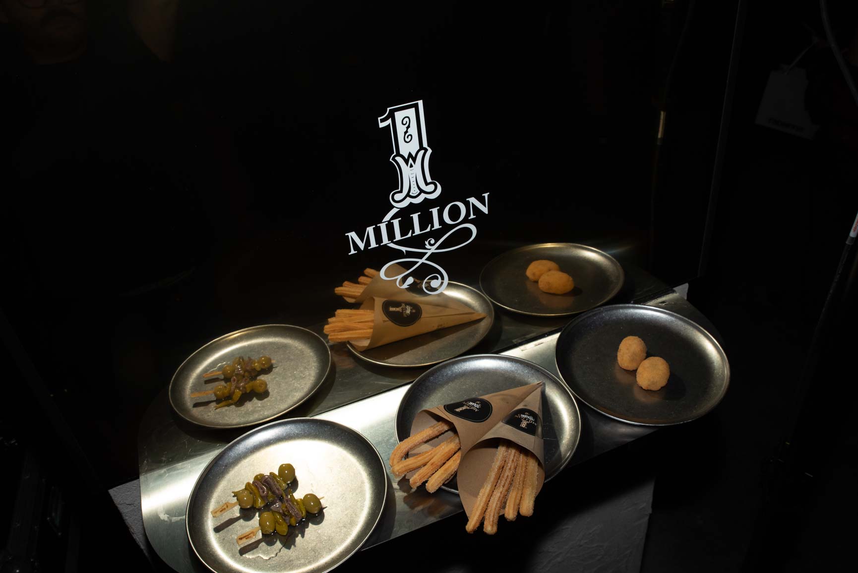 Catering con platos de metal, brochetas de aceitunas, churros en conos Rabanne y croquetas, fondo con logo de One Million, pop-up Madrid