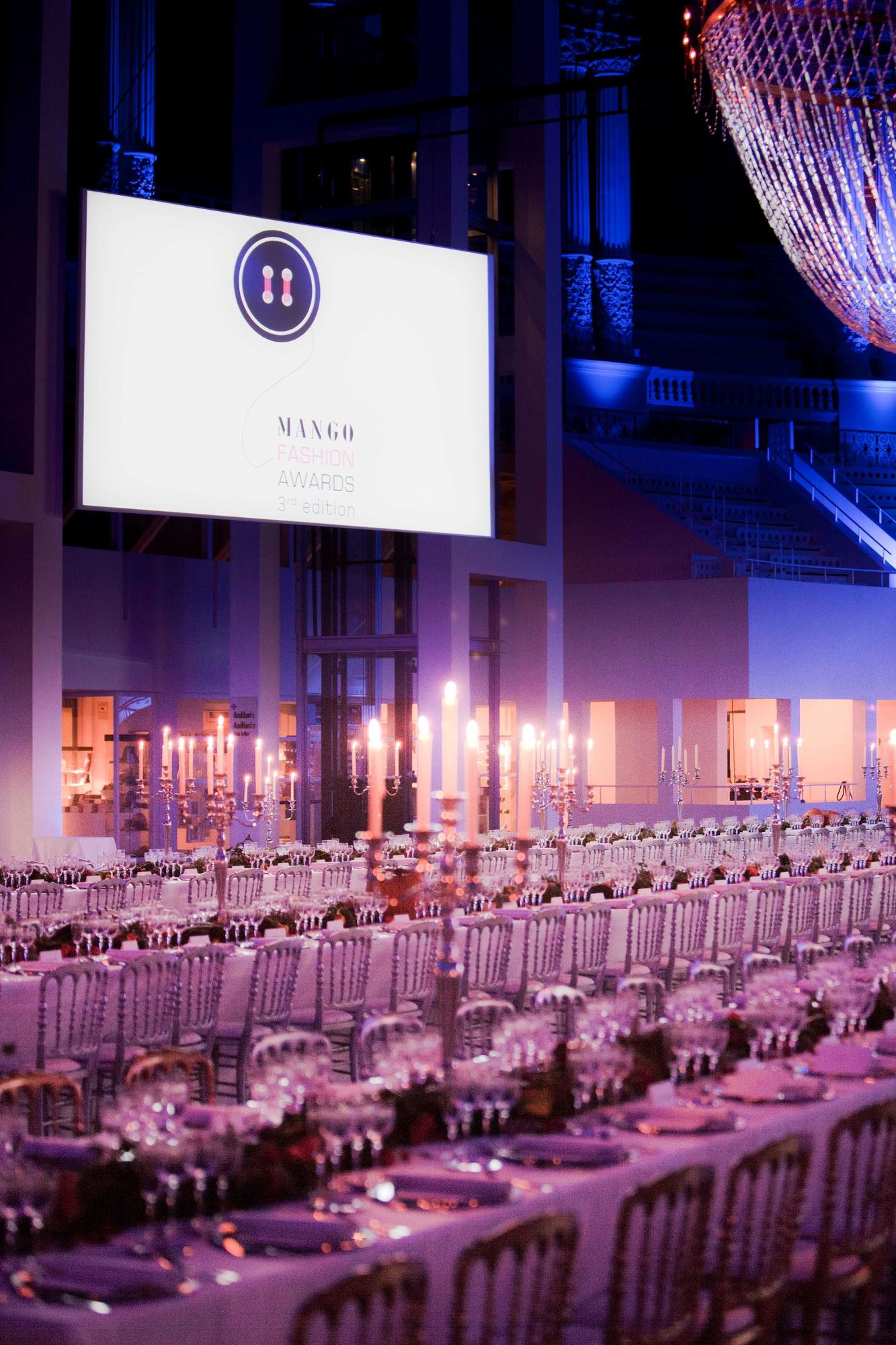 Larga mesa de gala con velas y lámparas de araña bajo iluminación violeta, y pantalla con el logotipo de Mango Fashion Awards, evento producido por ESMA Events