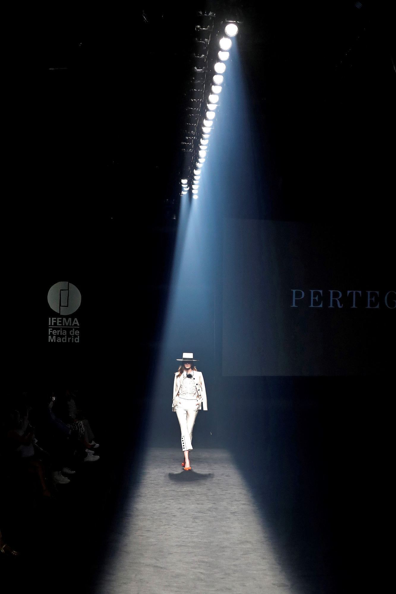 Modelo con traje blanco y sombrero caminando por una pasarela oscura bajo un haz de luz, en Mercedes-Benz Fashion Week Madrid SS 2021, producida por ESMA Events