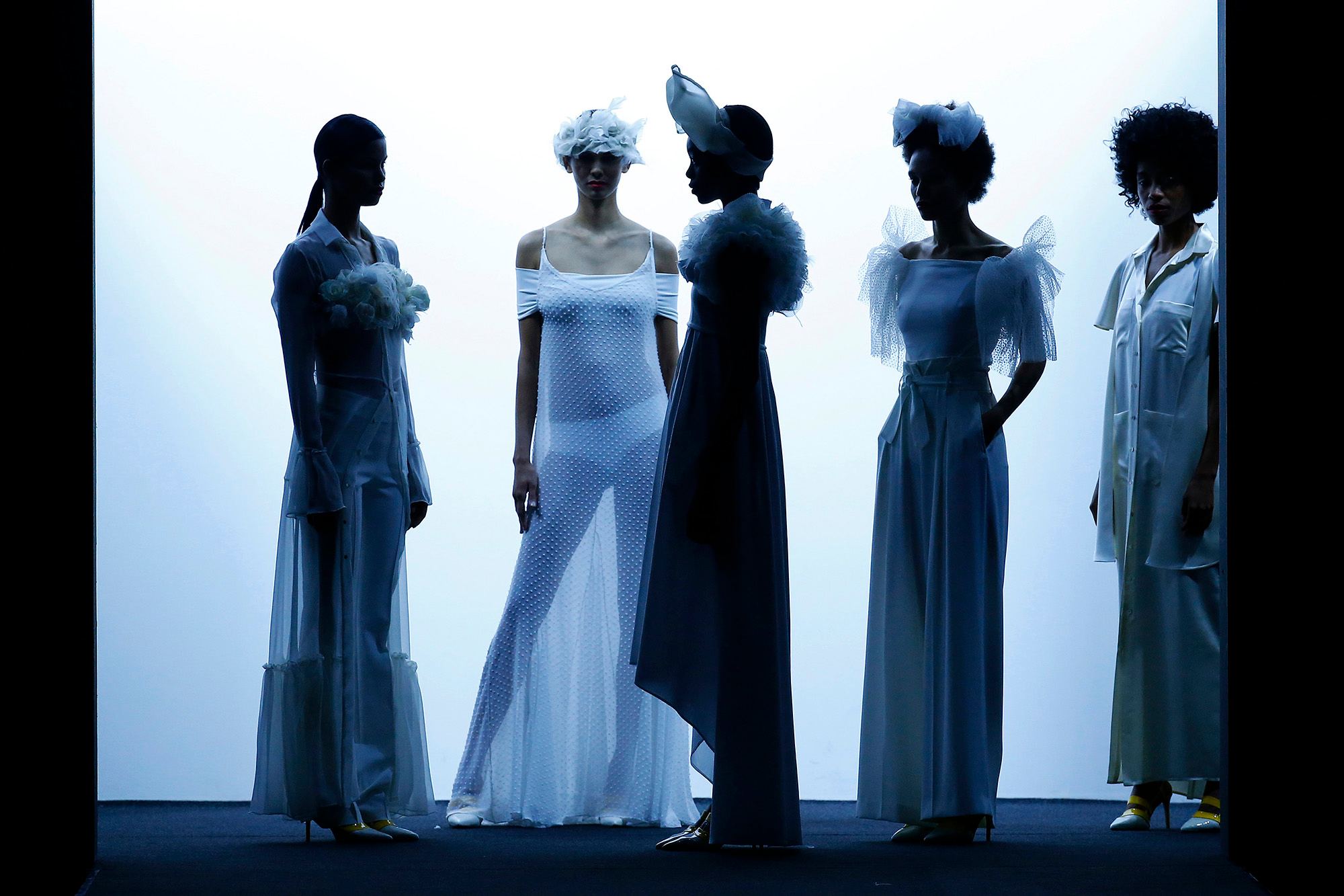 Cinco modelos a contraluz con vestidos azules y blancos y tocados, en Mercedes-Benz Fashion Week Madrid SS 2019, producida por ESMA Events