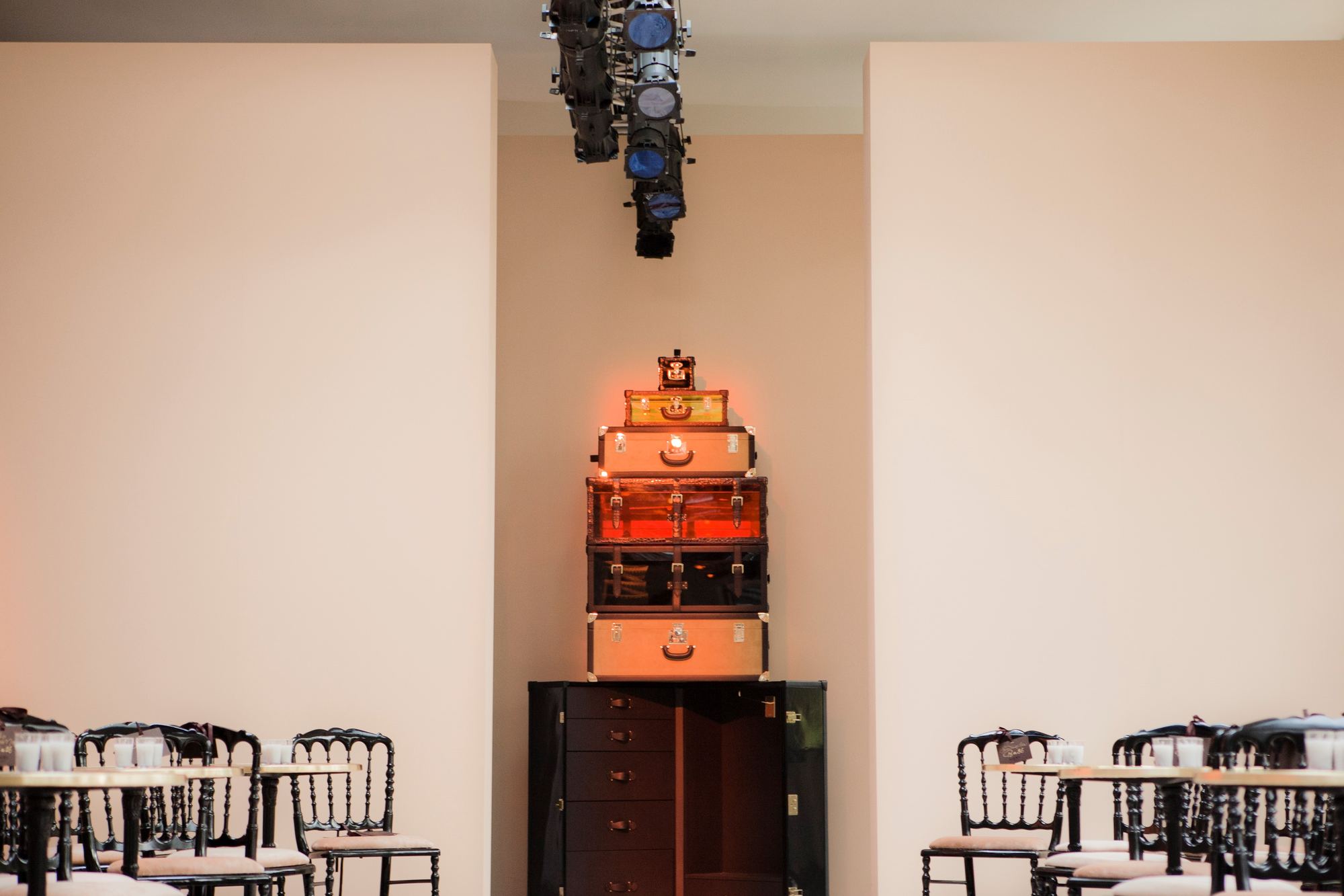 Maletas apiladas e iluminadas sobre un mueble de madera oscura, con sillas negras y focos de escenario, evento de Loewe producido por ESMA Events
