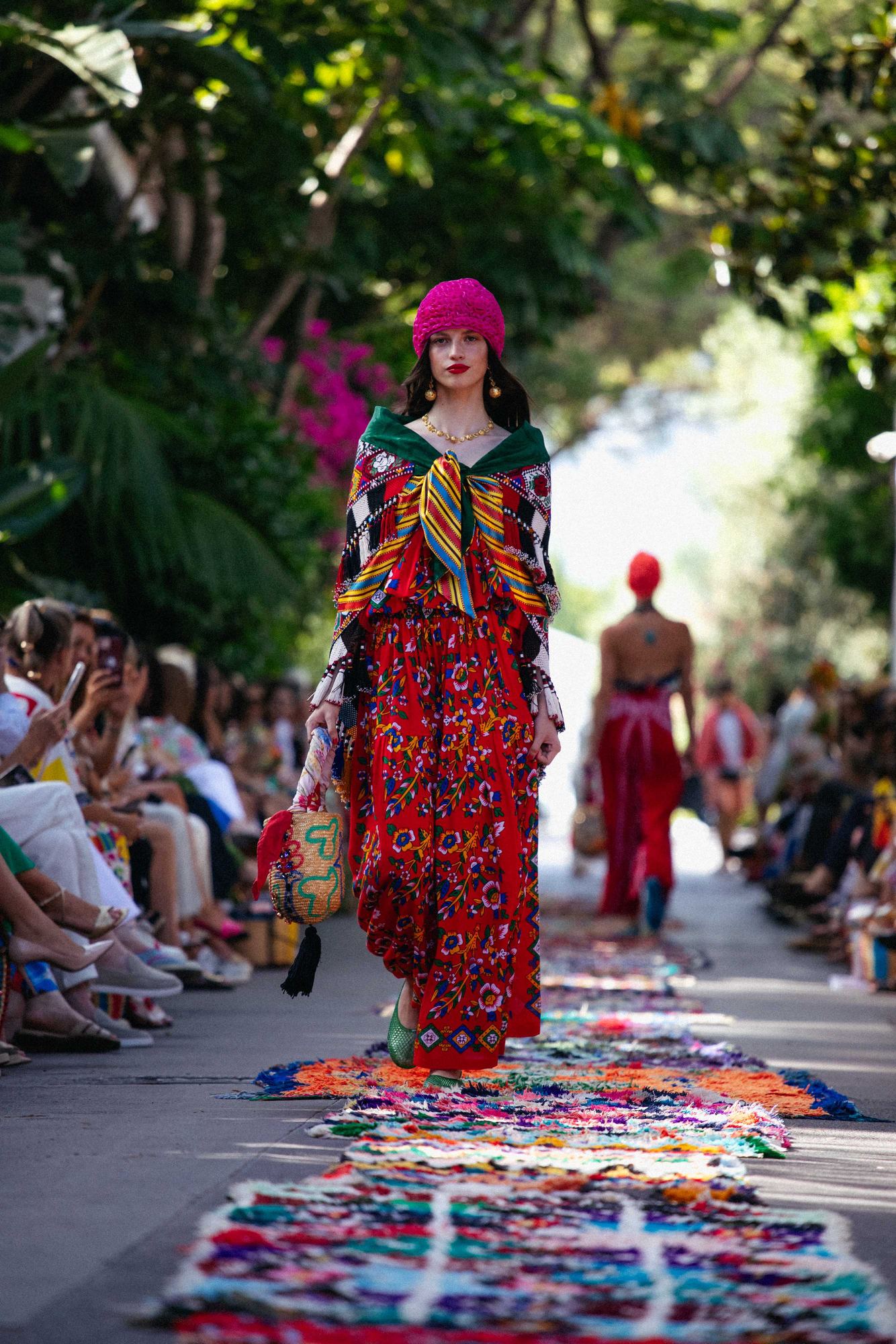 Modelo con look de colores intensos y gorro fucsia desfilando al aire libre entre vegetación sobre una alfombra de tejidos, desfile de Rianna + Nina producido por ESMA Events