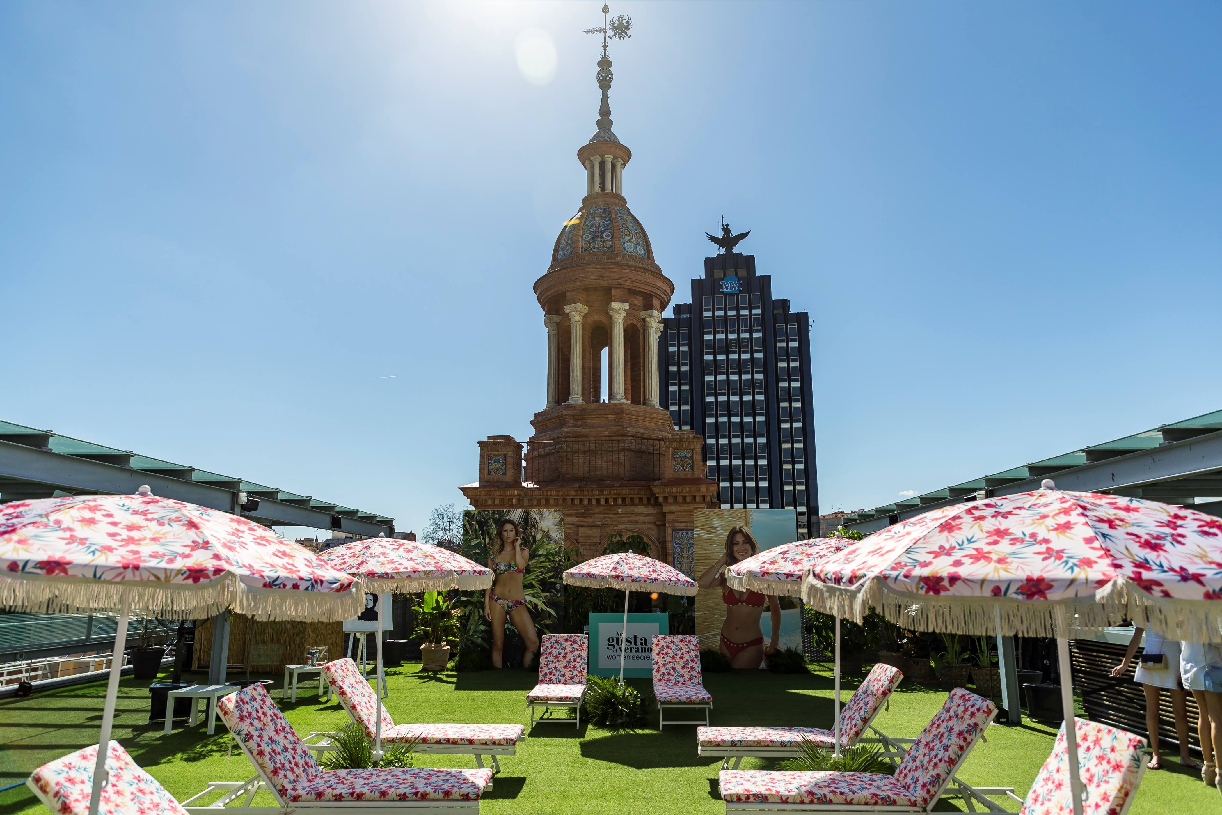 Terraza con césped, sombrillas de flores con flecos, tumbonas y una torre histórica al fondo, evento Women'secret Nos gusta el verano producido por ESMA Events
