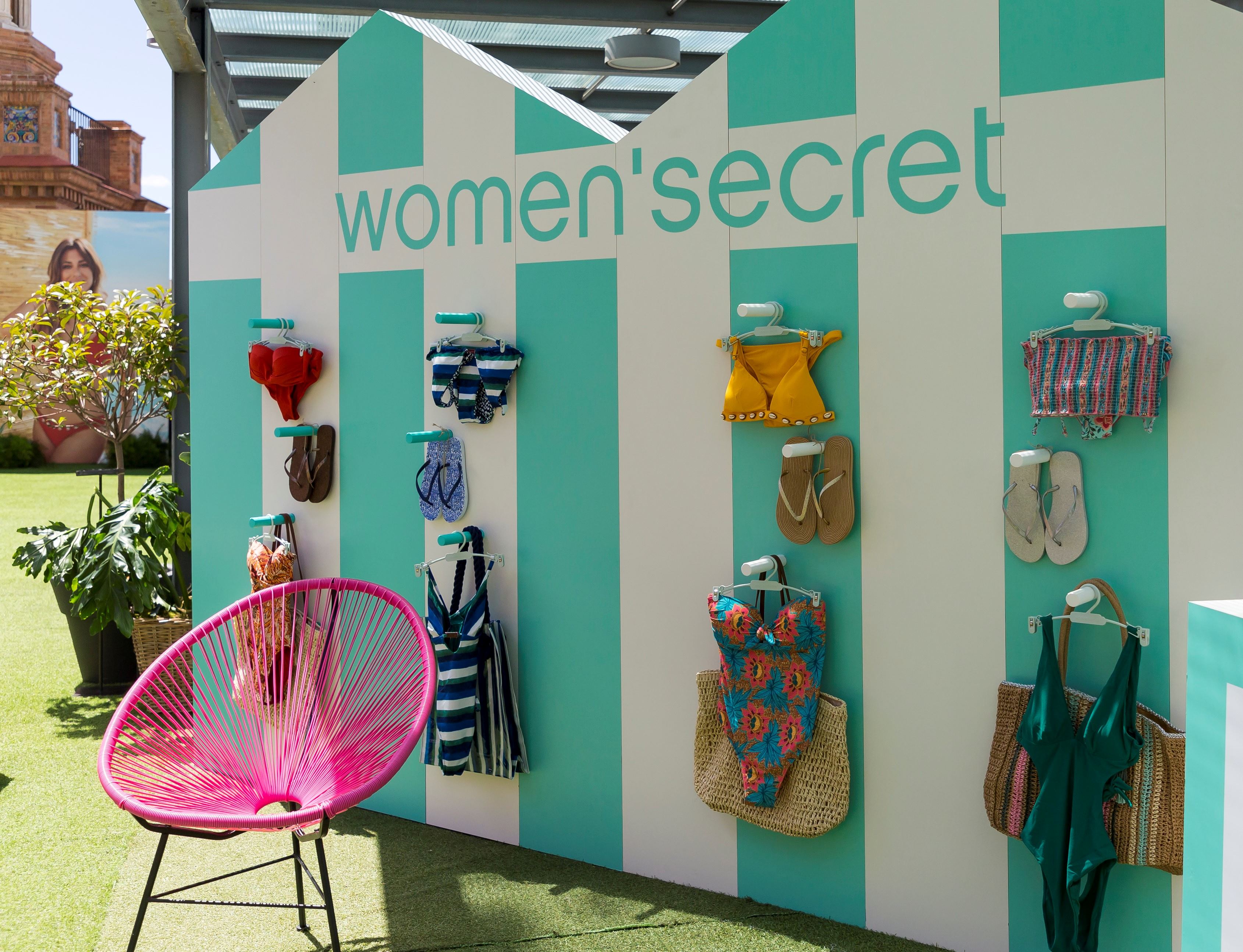 Pared de rayas turquesas y blancas con el logotipo de Women'secret y bañadores colgados, junto a una silla rosa, evento Women'secret Nos gusta el verano producido por ESMA Events