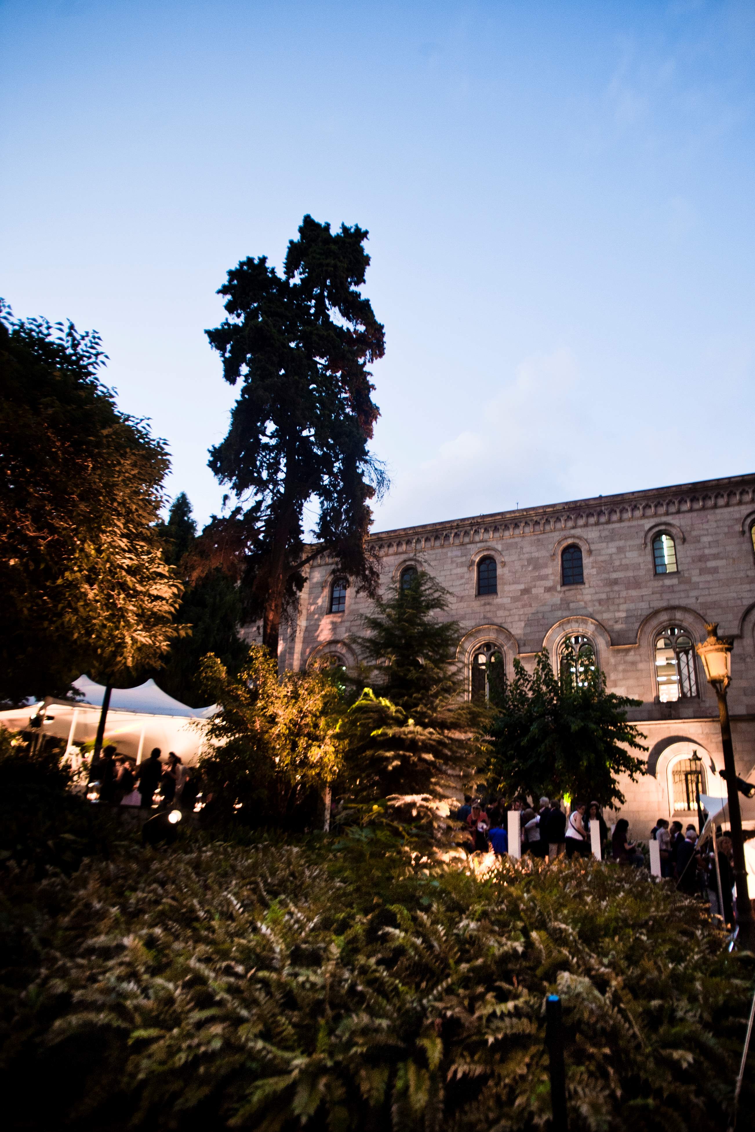 Jardín con árboles y un edificio histórico al fondo, con carpas blancas y invitados al atardecer, cocktail party de Mango producida por ESMA Events
