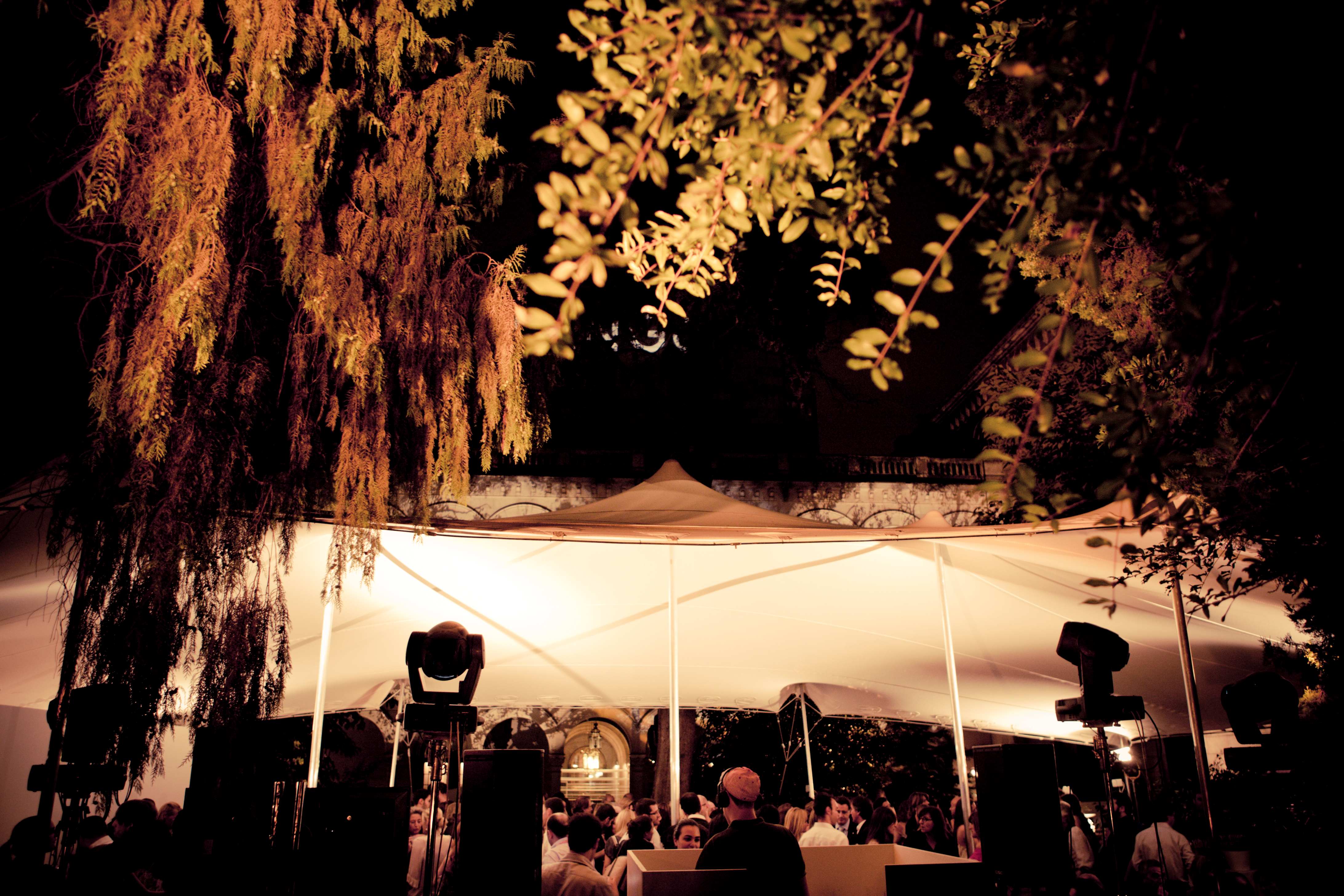 Carpa blanca iluminada entre árboles con focos móviles y público por la noche, cocktail party de Mango producida por ESMA Events