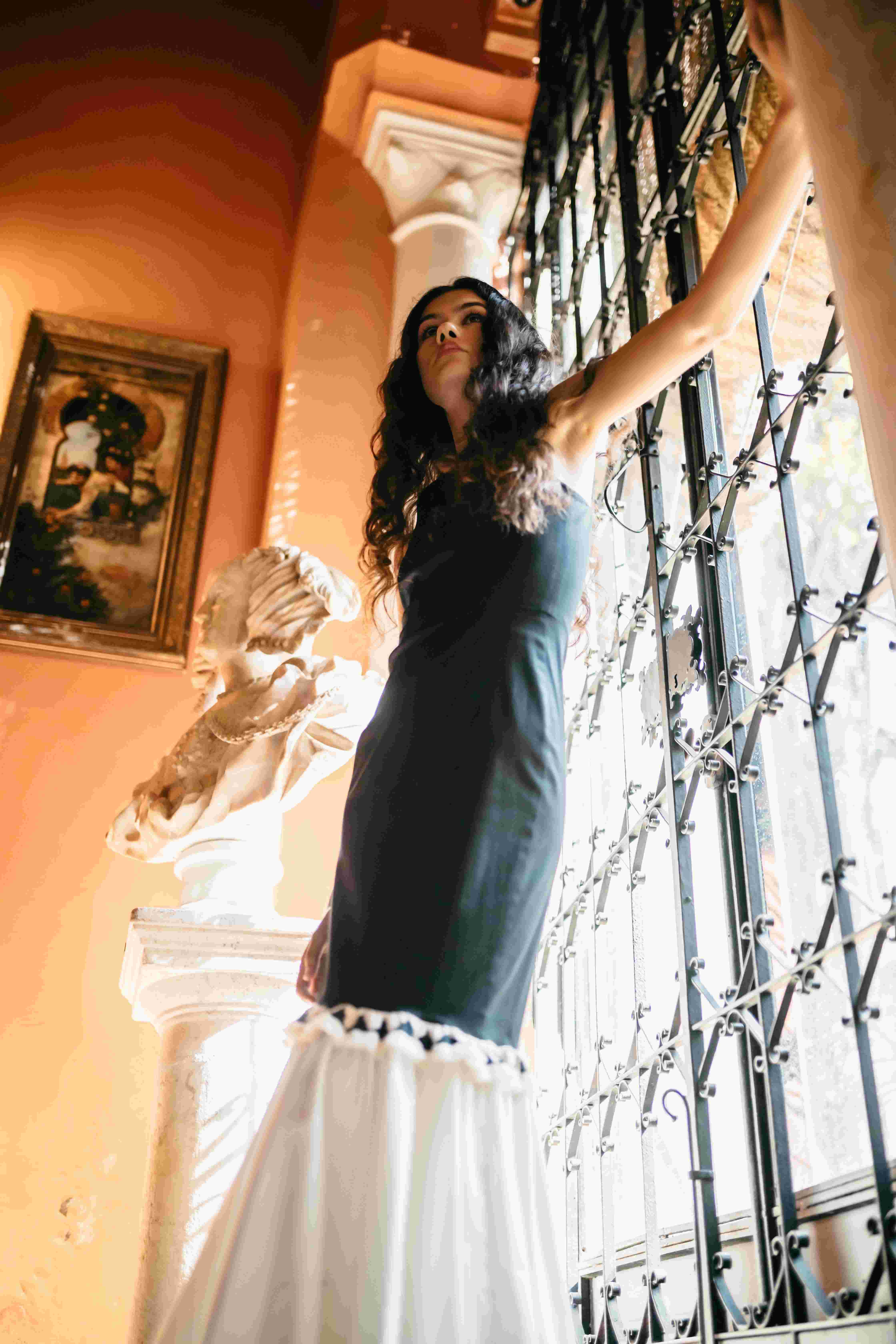 Modelo con vestido azul oscuro y falda blanca asomada a una reja de hierro forjado, desfile IQ Collection producido por ESMA Events