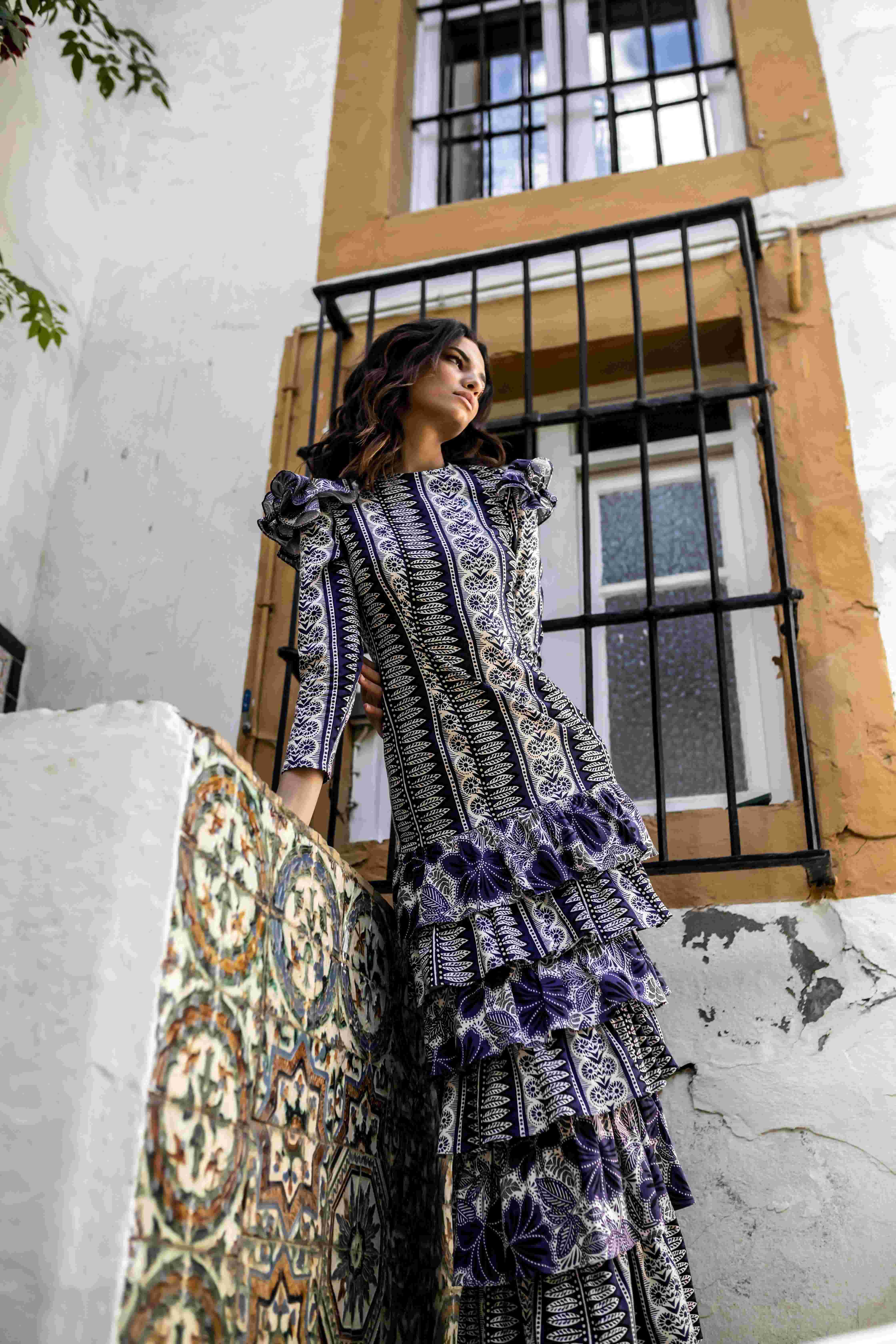 Modelo con vestido largo de volantes morados junto a una ventana enrejada y un azulejo, desfile IQ Collection producido por ESMA Events