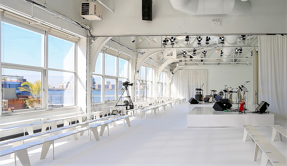 Sala blanca con ventanales sobre el río, bancos corridos para el público y banda al fondo, desfile Delpozo Nueva York SS 2018 producido por ESMA Events