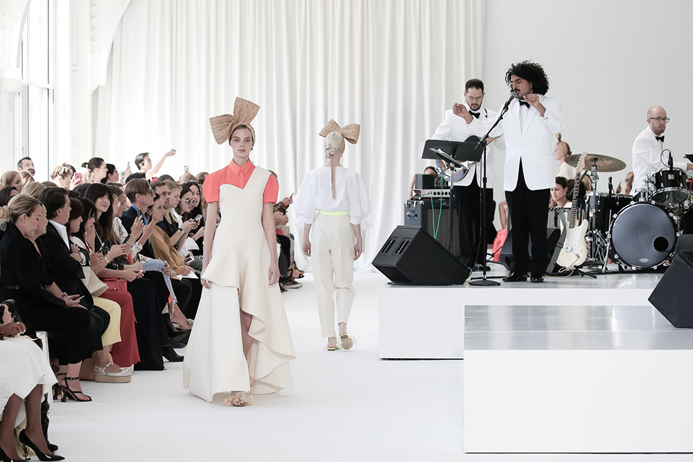Modelo con vestido blanco asimétrico caminando ante el público y la banda, desfile Delpozo Nueva York SS 2018 producido por ESMA Events