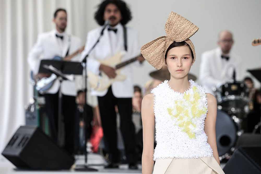 Modelo con top blanco de aplicaciones amarillas y lazo de rafia ante músicos de esmoquin blanco, desfile Delpozo Nueva York SS 2018 producido por ESMA Events