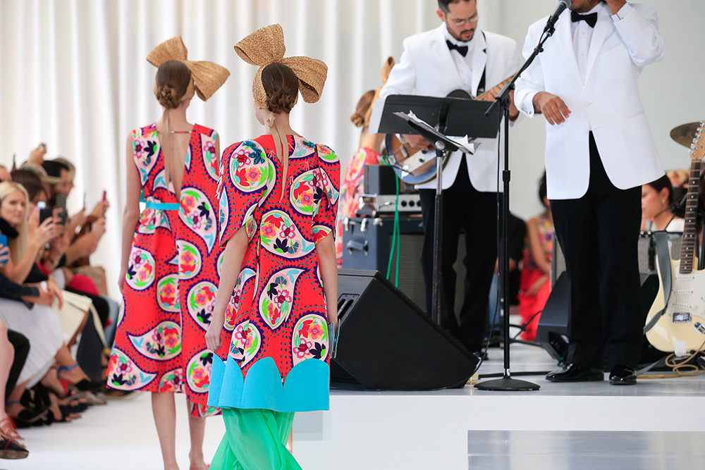 Dos modelos de espaldas con vestidos estampados rojos y lazos de rafia ante la banda, desfile Delpozo Nueva York SS 2018 producido por ESMA Events