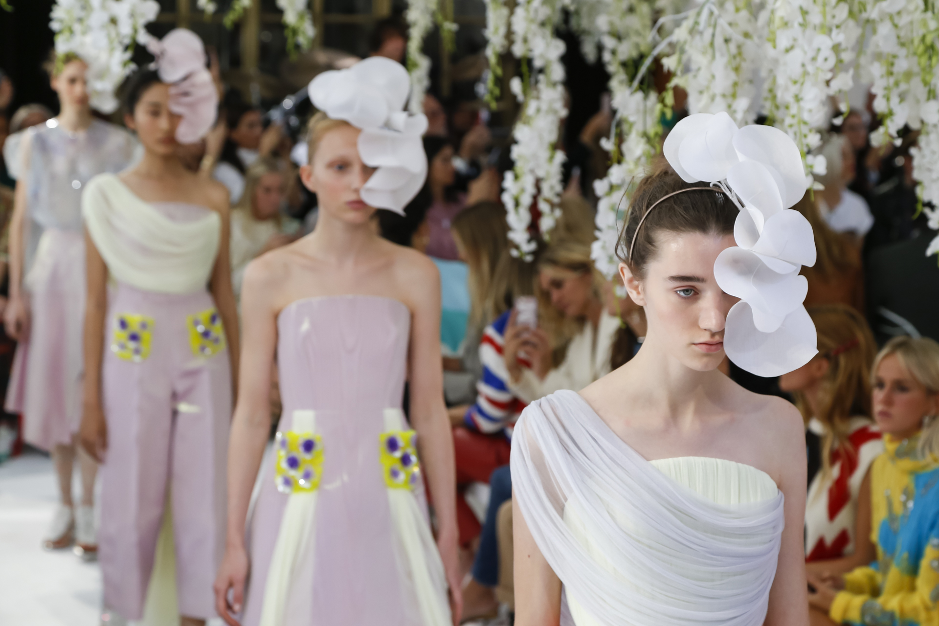 Tres modelos con tocados blancos de pétalos y prendas en lila y blanco caminando ante el público, desfile Delpozo Londres SS 2019 producido por ESMA Events