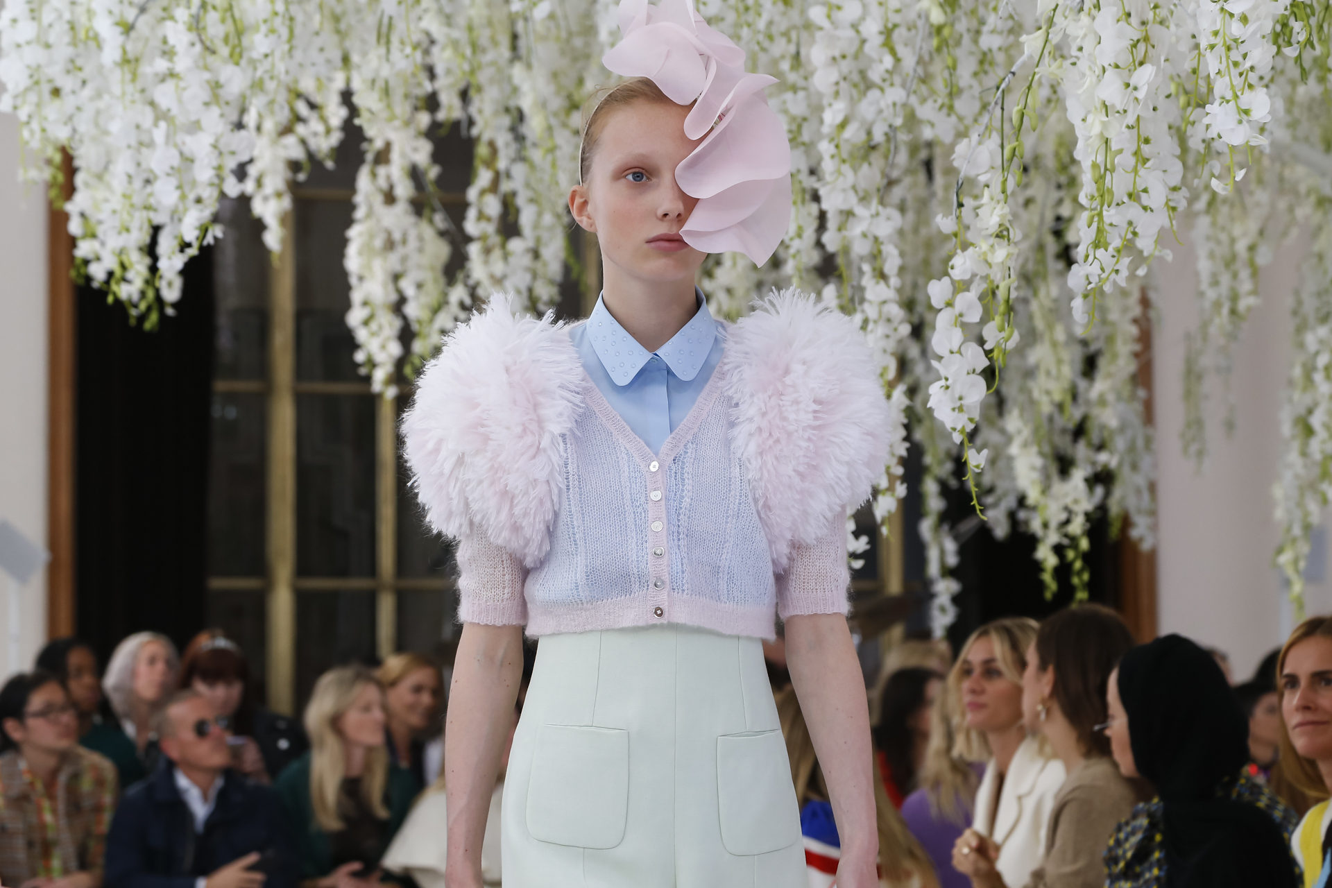 Primer plano de una modelo con tocado rosa y chaqueta de plumas rosas bajo glicinias colgantes, desfile Delpozo Londres SS 2019 producido por ESMA Events