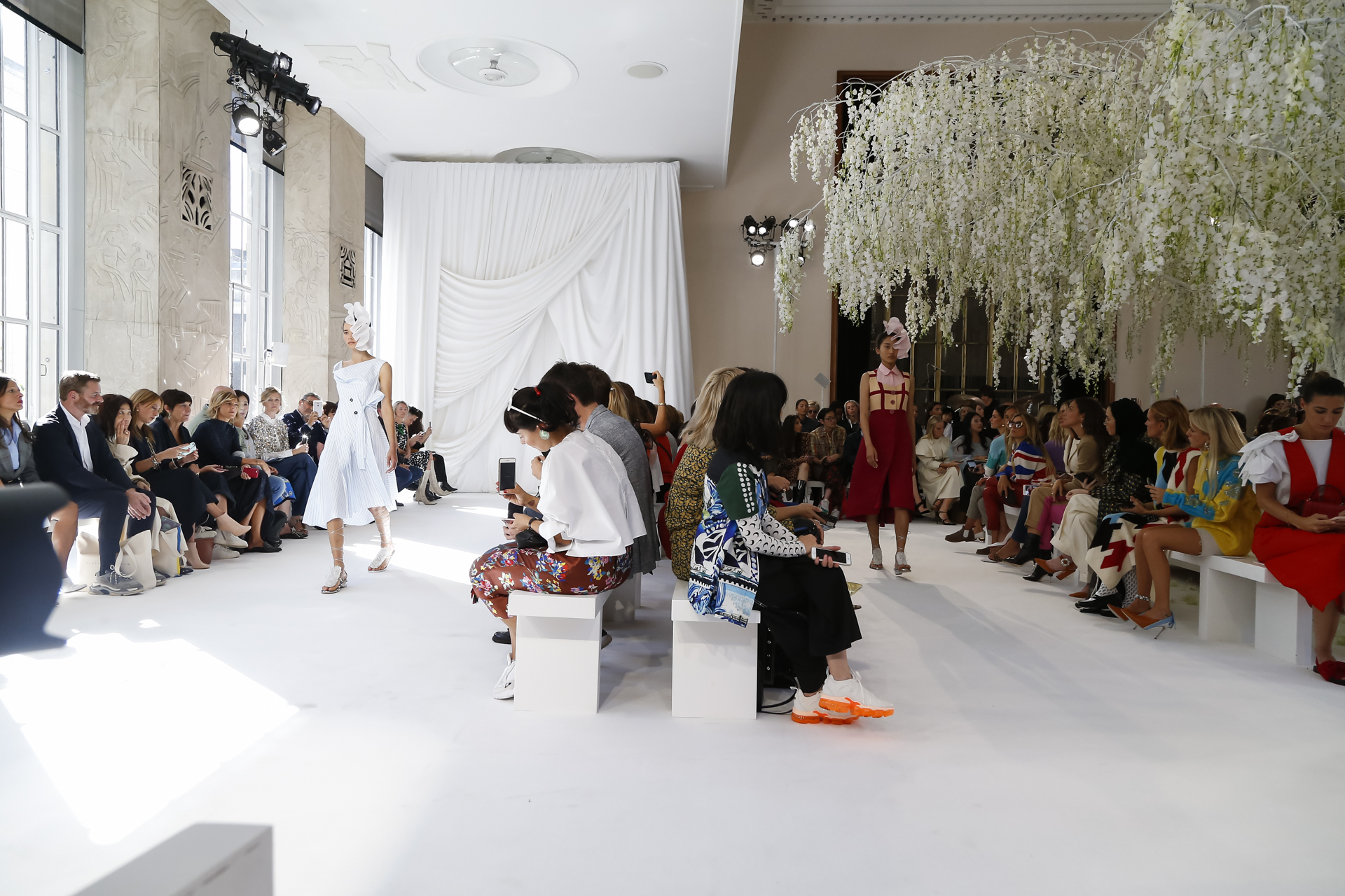 Modelo con vestido blanco en la pasarela blanca, con público a ambos lados y glicinias al fondo, desfile Delpozo Londres SS 2019 producido por ESMA Events